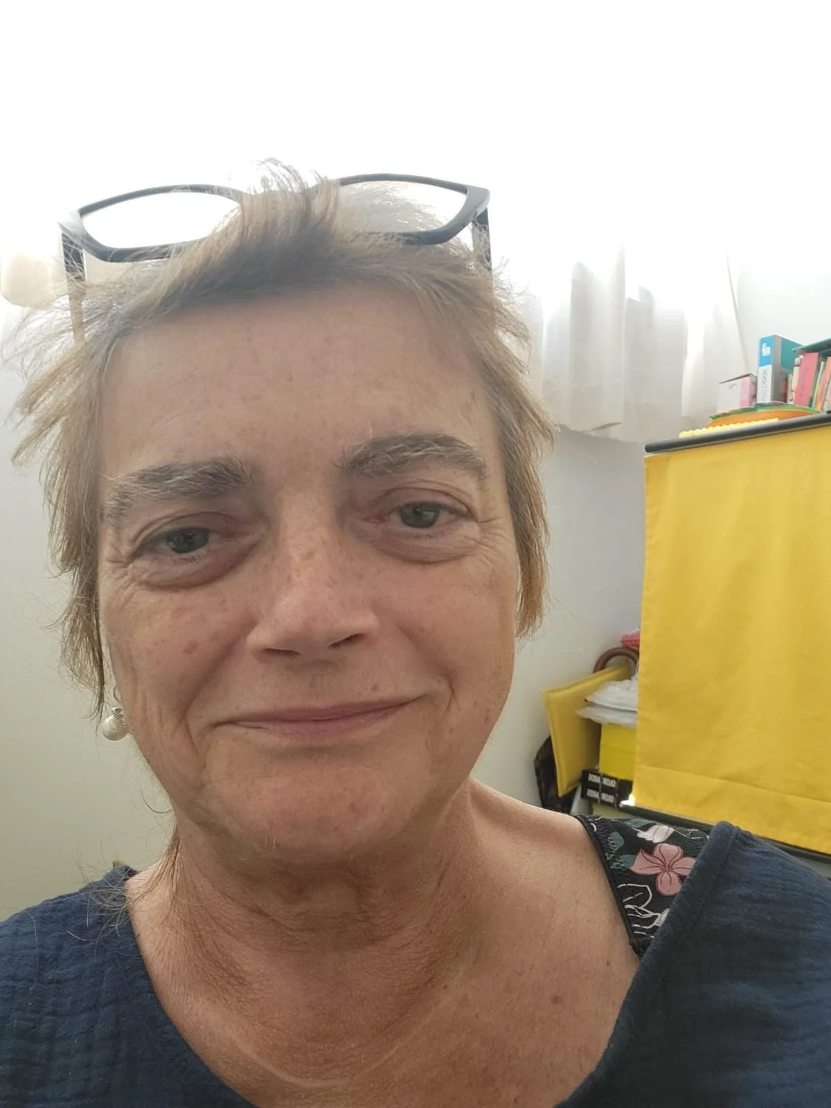 Primissimo piano di una donna con capelli corti e castani, occhiali poggiati sulla testa, sorriso leggero, sfondo di una stanza con scaffali e scatole di vari colori.