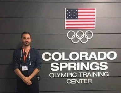 Persona che si trova davanti a un muro con lo stemma delle Olimpiadi, la bandiera degli Stati Uniti e le scritte "Colorado Springs Olympic Training Center".