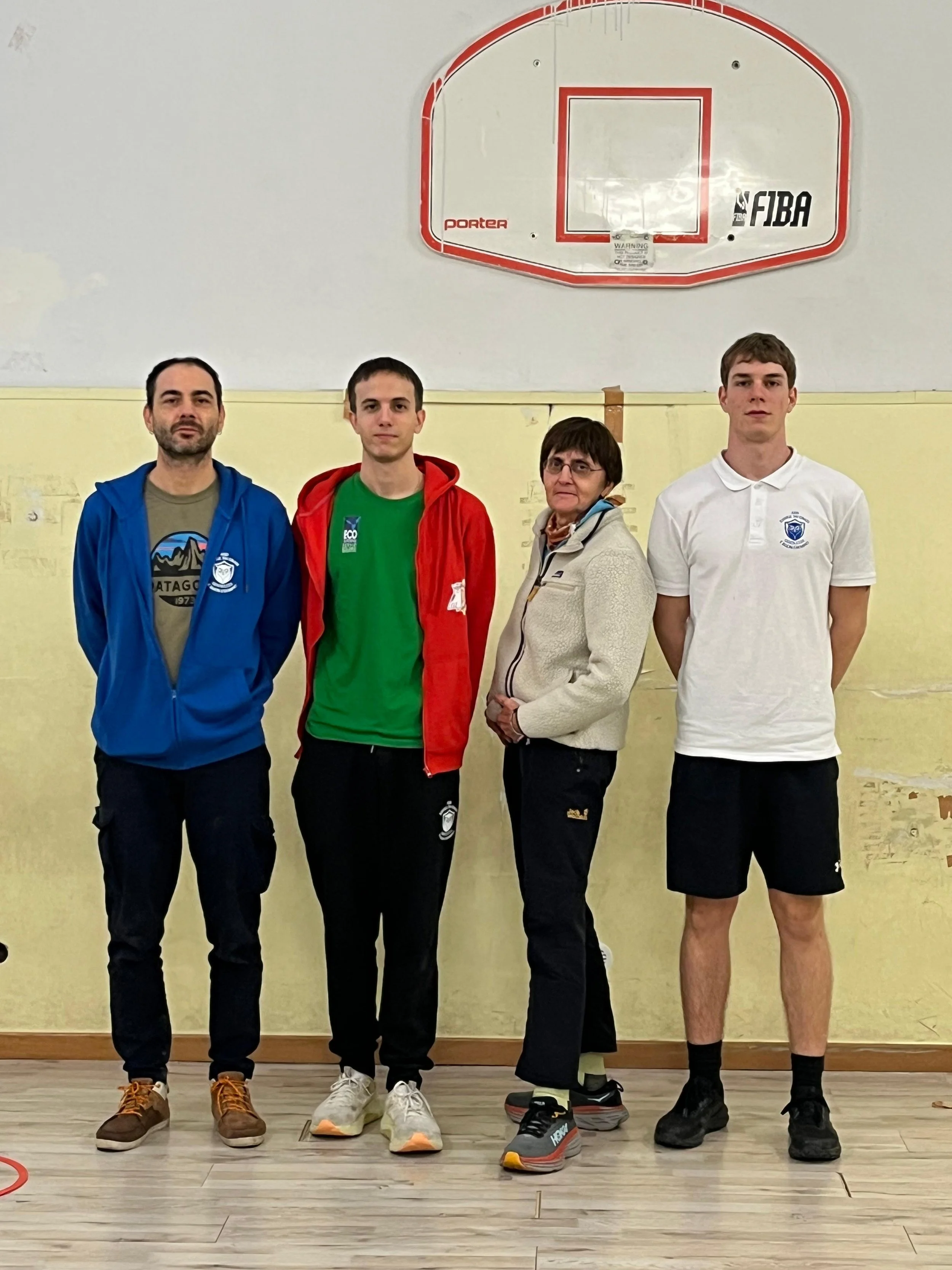 Quattro persone in piedi davanti a un muro giallo chiaro con un canestro da basket appeso sopra. Sono di fronte, tre uomini e una donna, indossano abbigliamento sportivo.