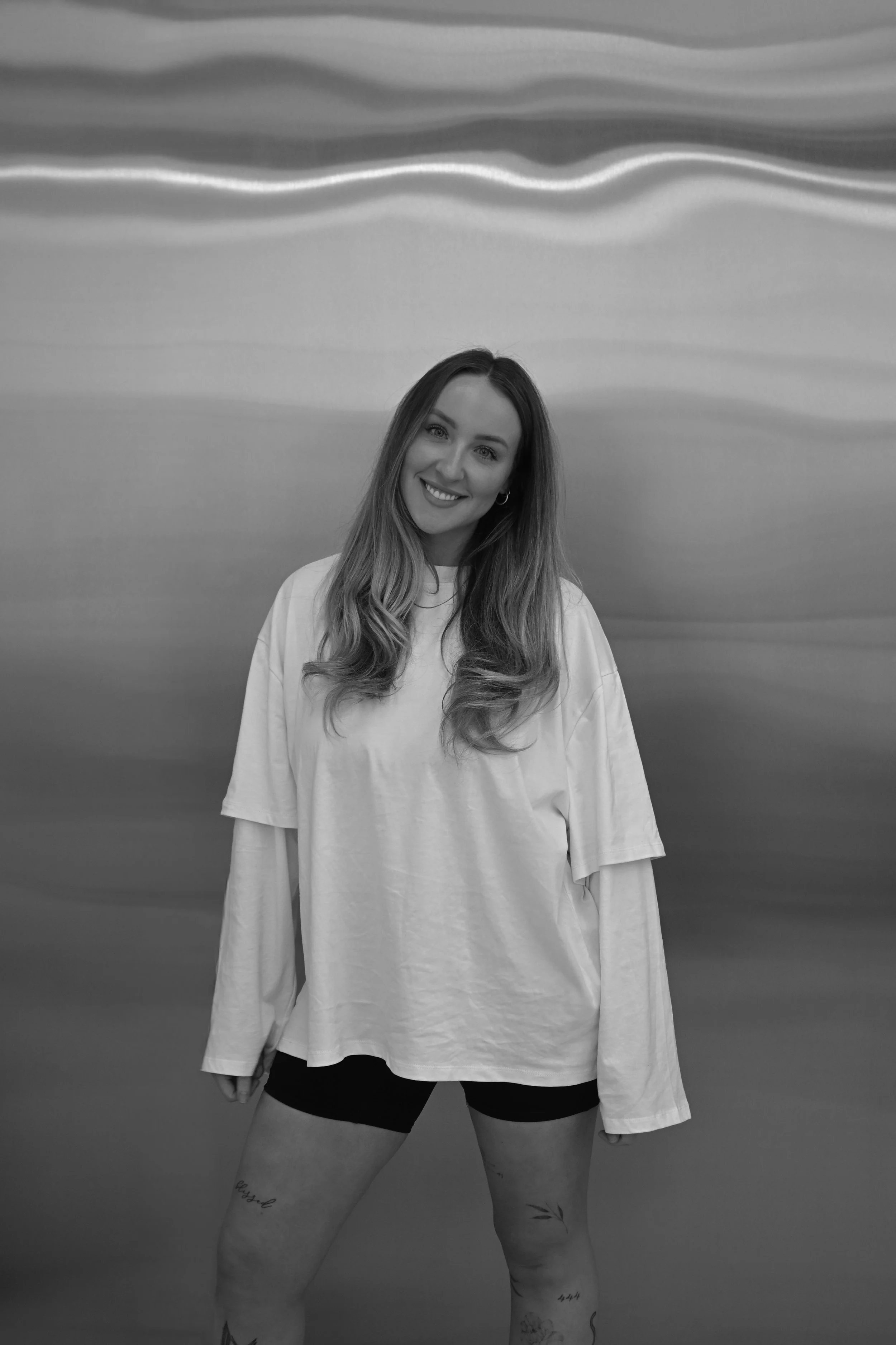 A young woman with long wavy hair posing against a metallic wavy background, smiling at the camera, wearing a loose long-sleeve shirt and shorts, with tattoos on her thighs.