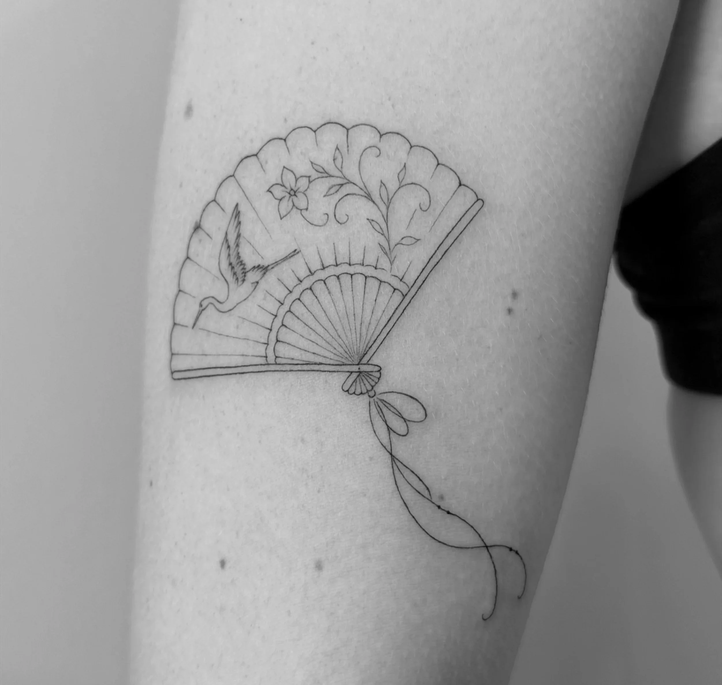 A black-and-white line drawing of a traditional folding fan with floral and vine motifs, and a tassel hanging from the base.