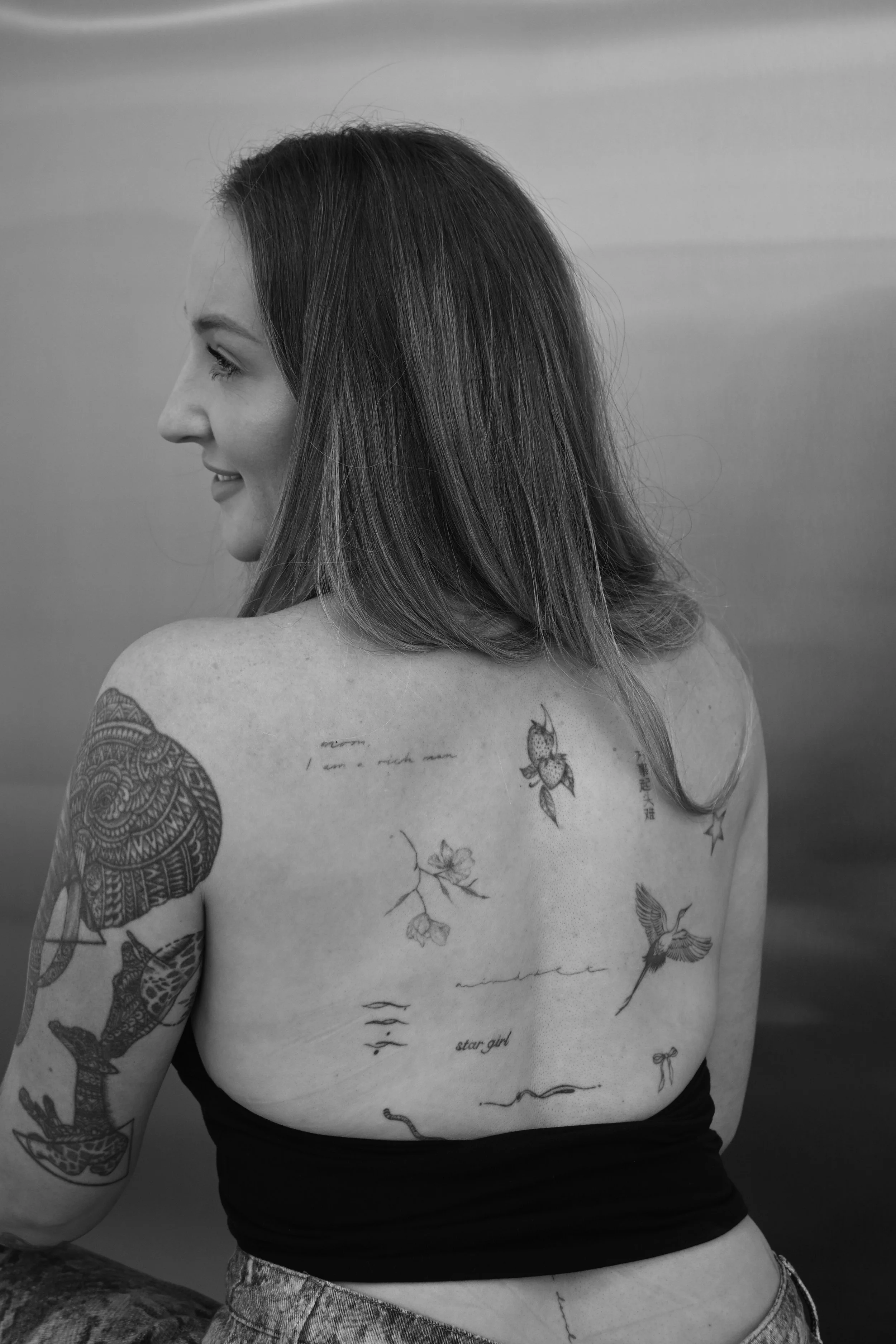 A woman with long, dark hair and tattoos on her shoulder and back, wearing a black top and patterned pants, stands against a plain background, looking to the side.