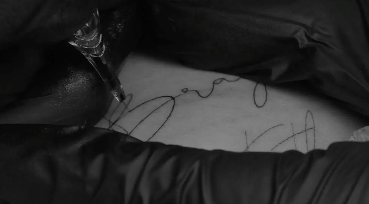 Person with black gloves tattooing a person's skin with a tattoo needle, creating a tattoo design.