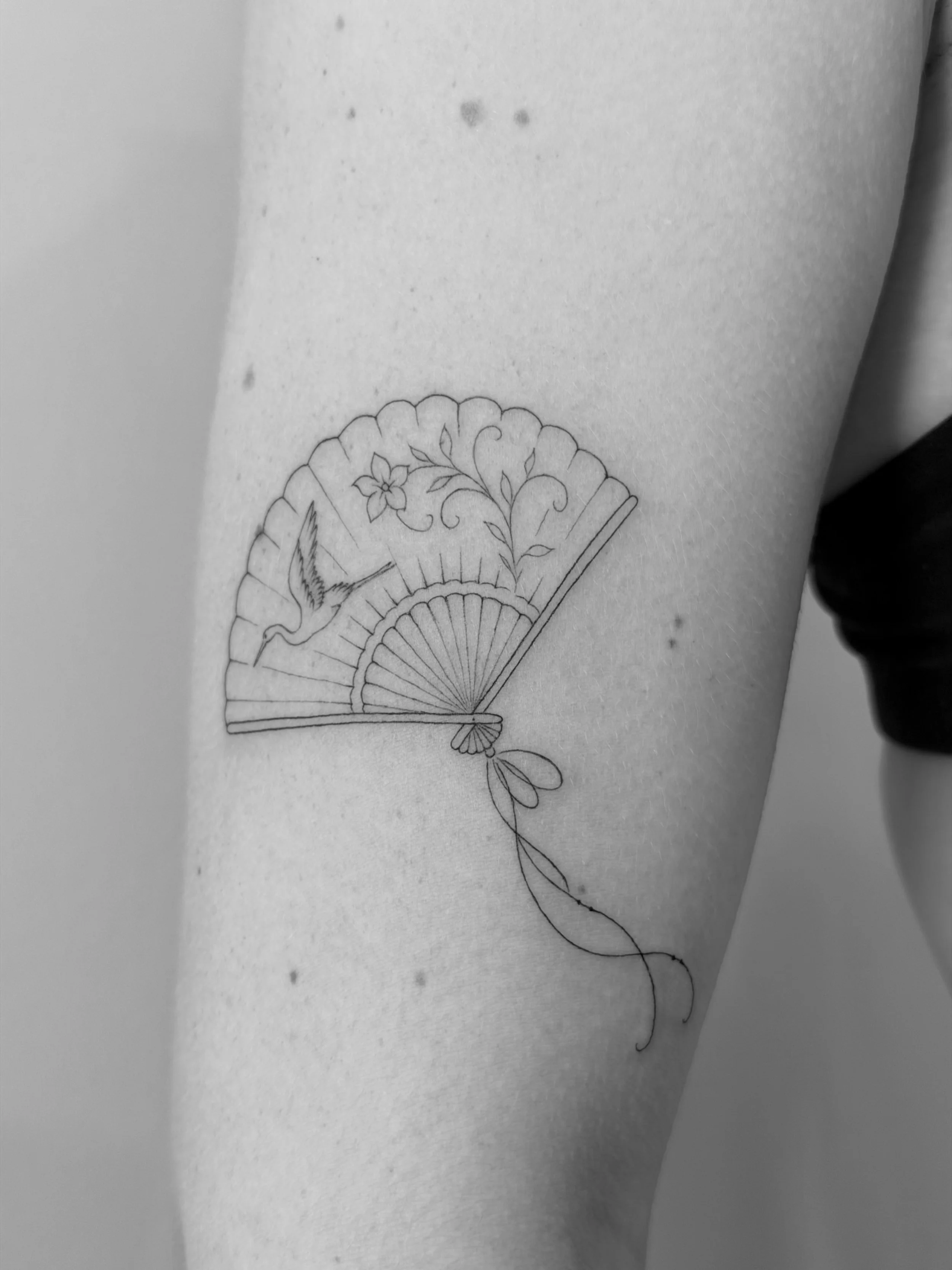 A black and white tattoo of a decorative hand fan with floral designs on a person's arm.