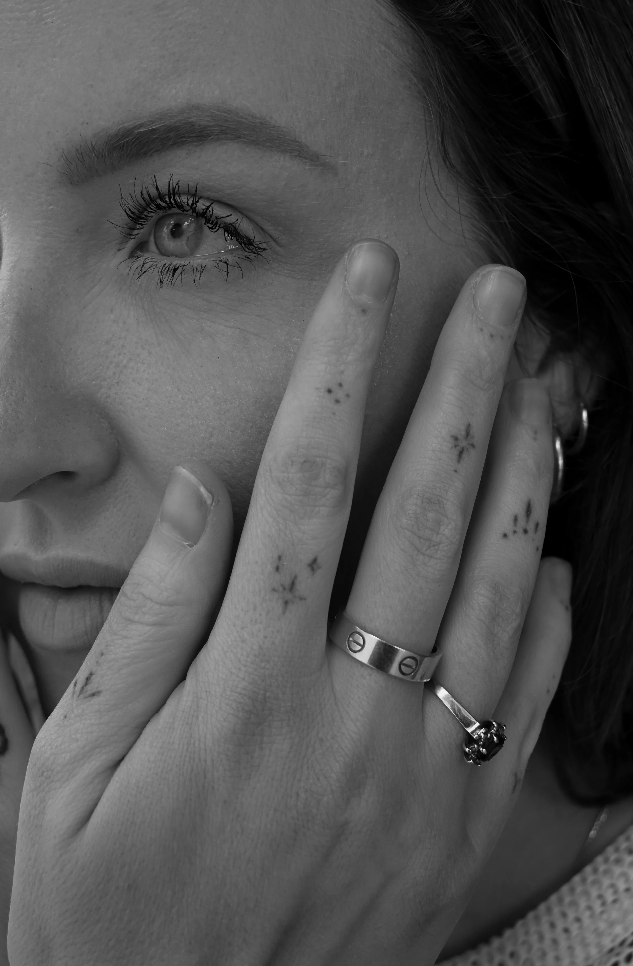 Close-up of a woman's face partially covered by her hand, which has small star tattoos on her fingers. She is wearing a ring and a bracelet with a skull charm. Her eye is visible, with long eyelashes and well-groomed eyebrows, and her hair is dark an