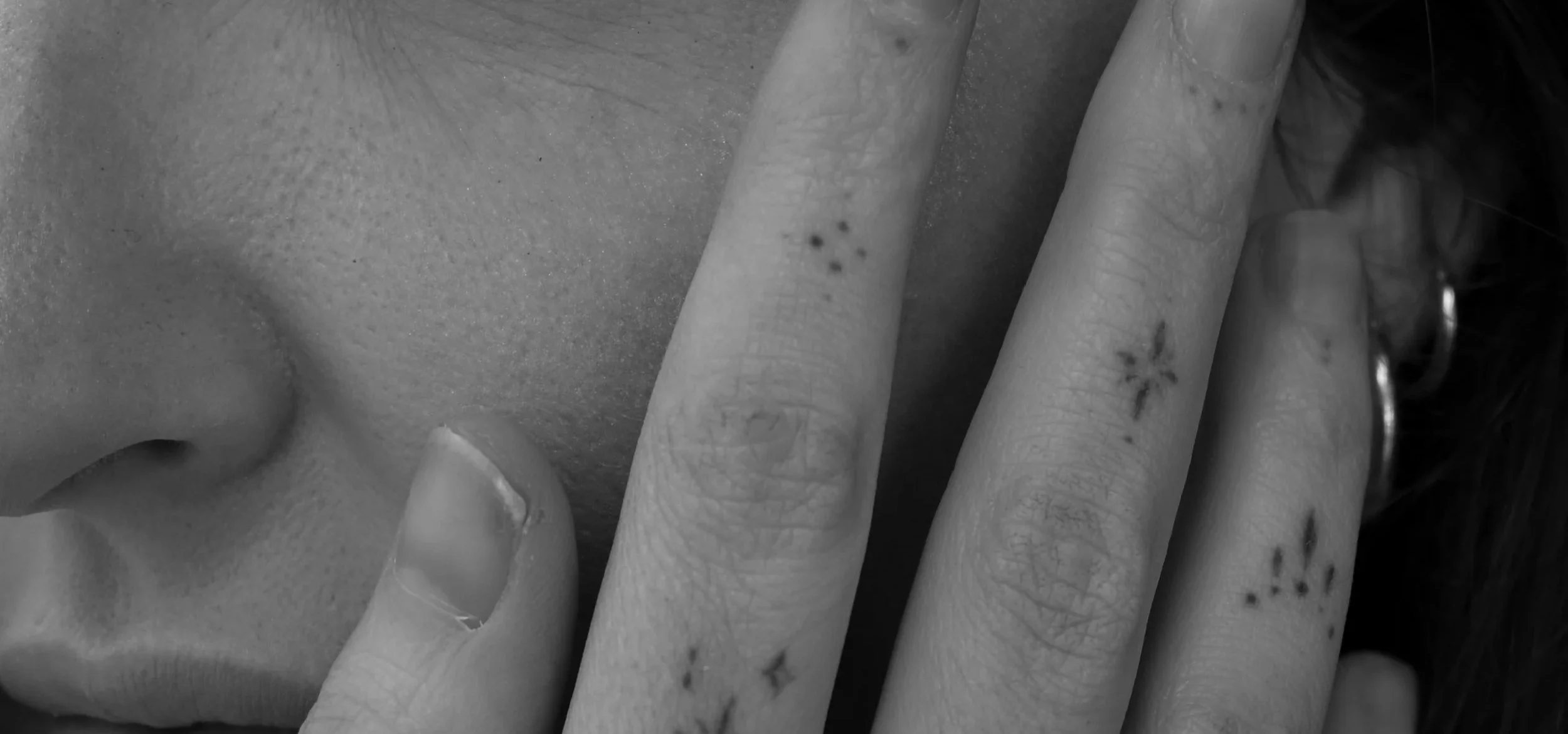 Close-up of a person's face and fingers, with the fingers covering part of the face, revealing small tattoos on the fingers.