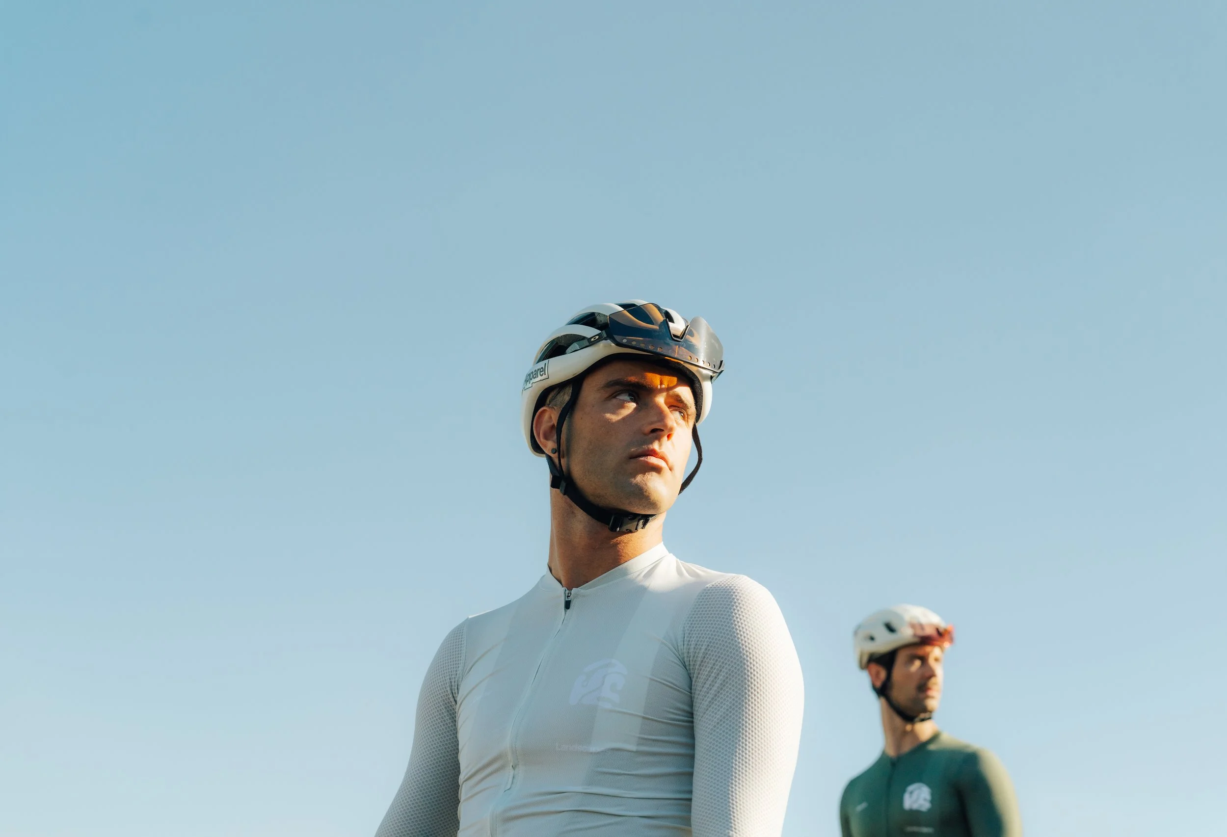 Two men wearing cycling helmets and aerodynamic clothing standing outdoors under a clear blue sky.