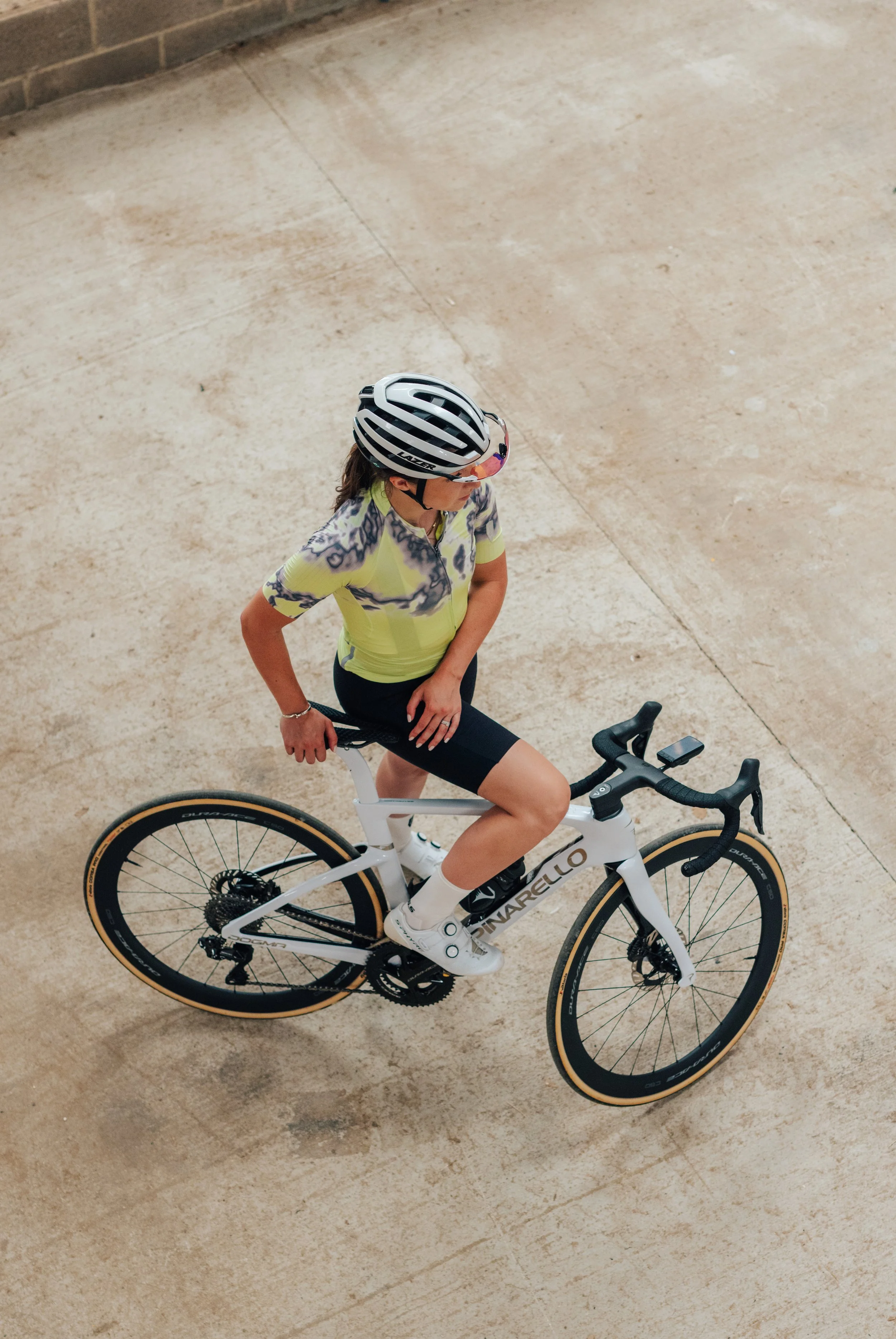 A woman wearing a helmet and cycling jersey, sitting on a white Pinarello bicycle on a beige concrete surface.