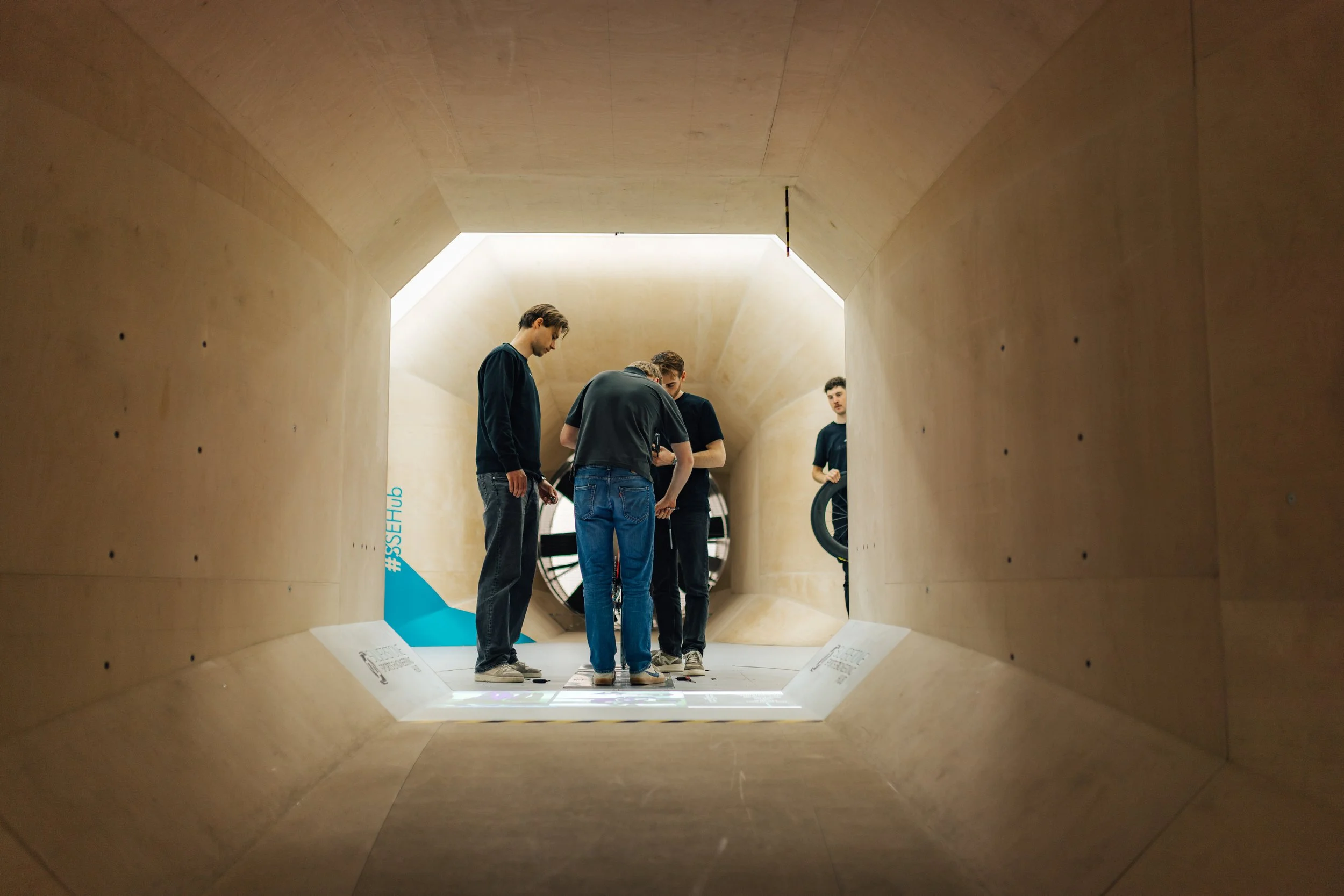 Four young men standing inside a wooden tunnel-like structure with lighting at the top and a large circular opening at the door. They are looking down at an object on the ground.
