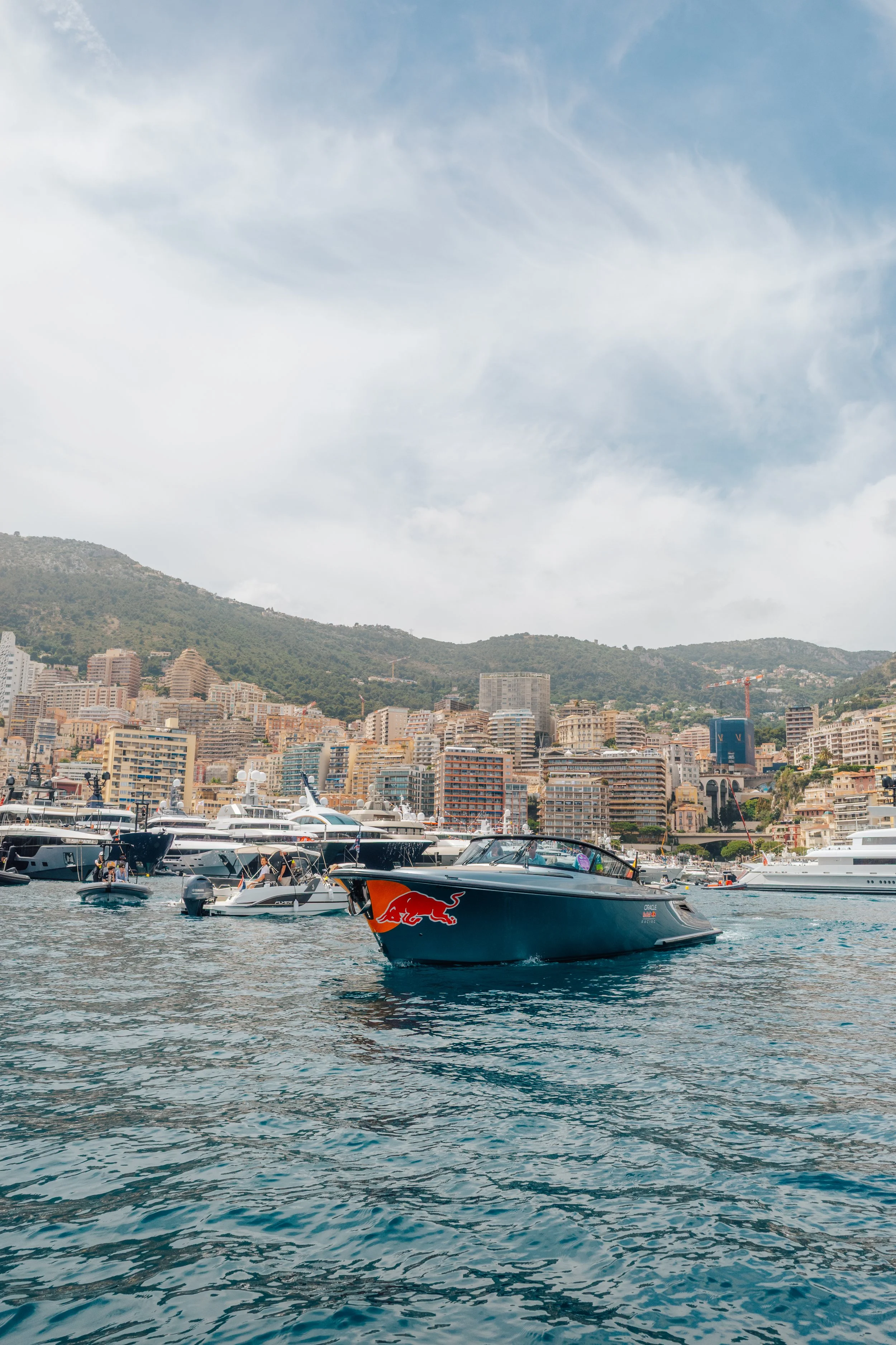 A marina filled with yachts and boats, with a city and hillside in the background. The sky is partly cloudy.