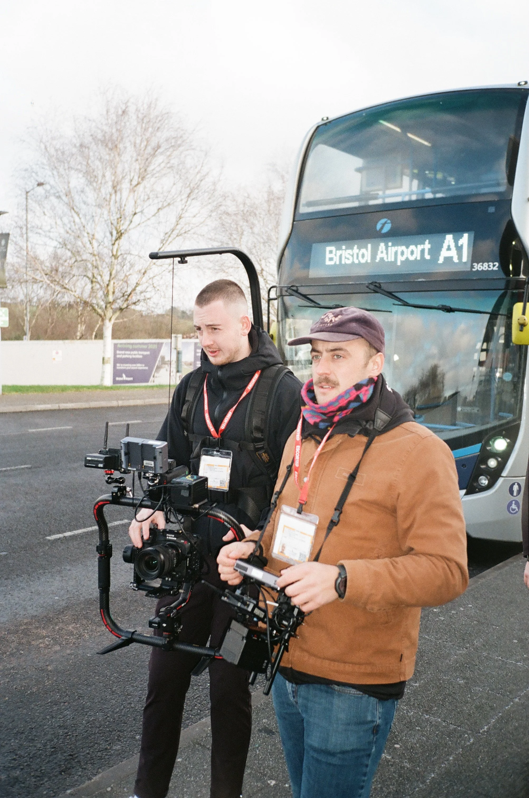 Two men holding professional camera equipment, standing outdoors in front of a double-decker bus labeled 'Bristol Airport A1,' with bare trees and parking lot in the background.