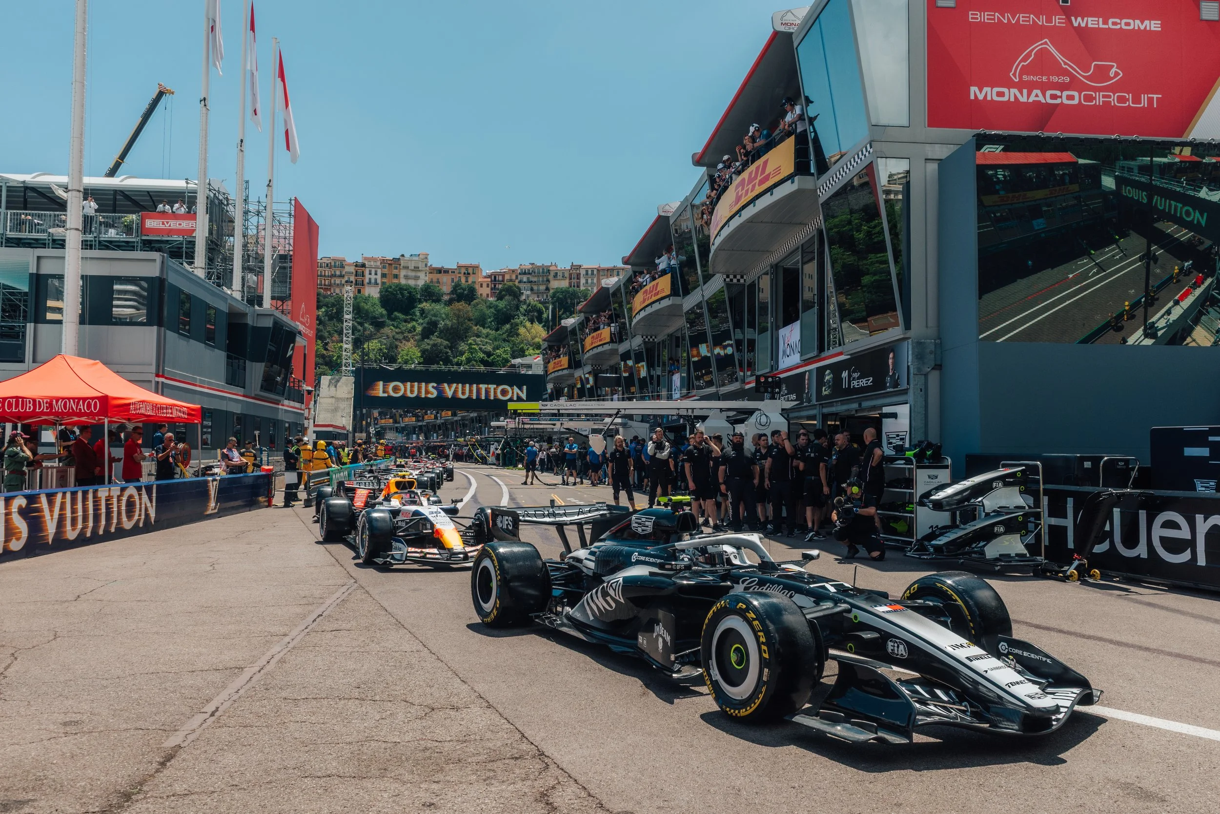 Formula 1 race cars in the pit lane of Monaco Grand Prix with team members and spectators, Monaco Circuit banners, and a scenic hillside background.