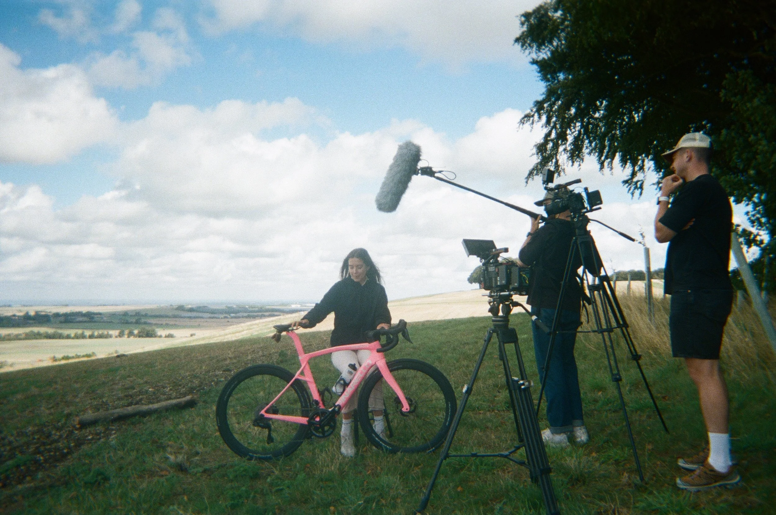 A filming crew setting up outdoors with a woman on a pink bicycle, a camera on a tripod, a person holding a microphone boom, and another person observing near a tree in a landscape with fields and parsley clouds.