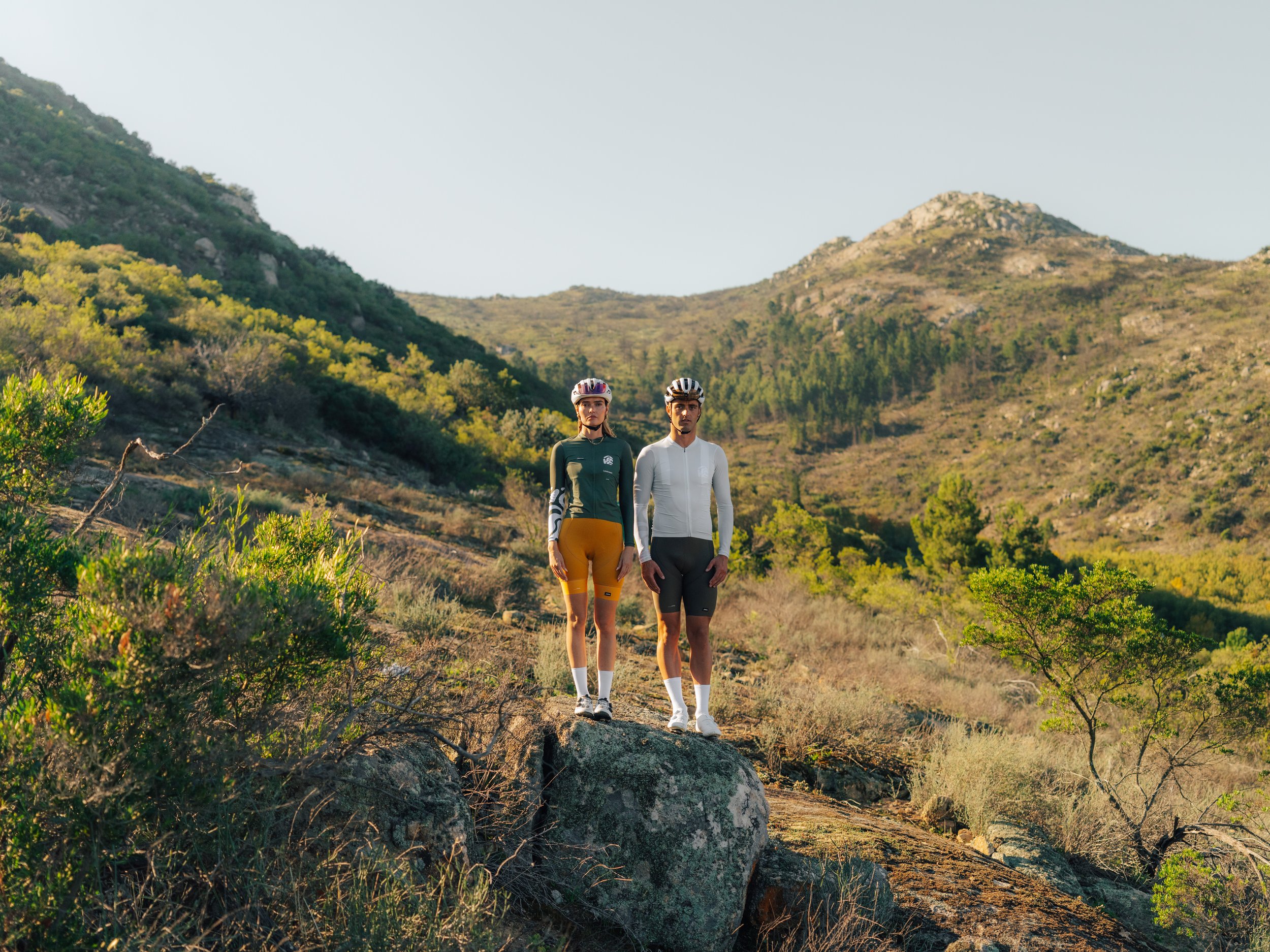 Two cyclists standing outdoors on rocky terrain with hills and sparse vegetation in the background, wearing helmets and athletic clothing.