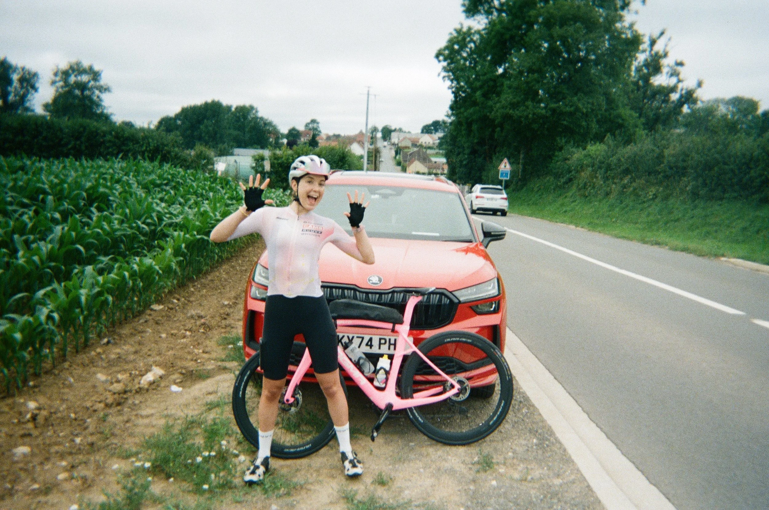 A young woman in cycling gear and a helmet standing on the roadside with a pink bicycle, smiling and waving. Behind her, a red car is parked, and the background includes a rural road, green fields, trees, and a cloudy sky.