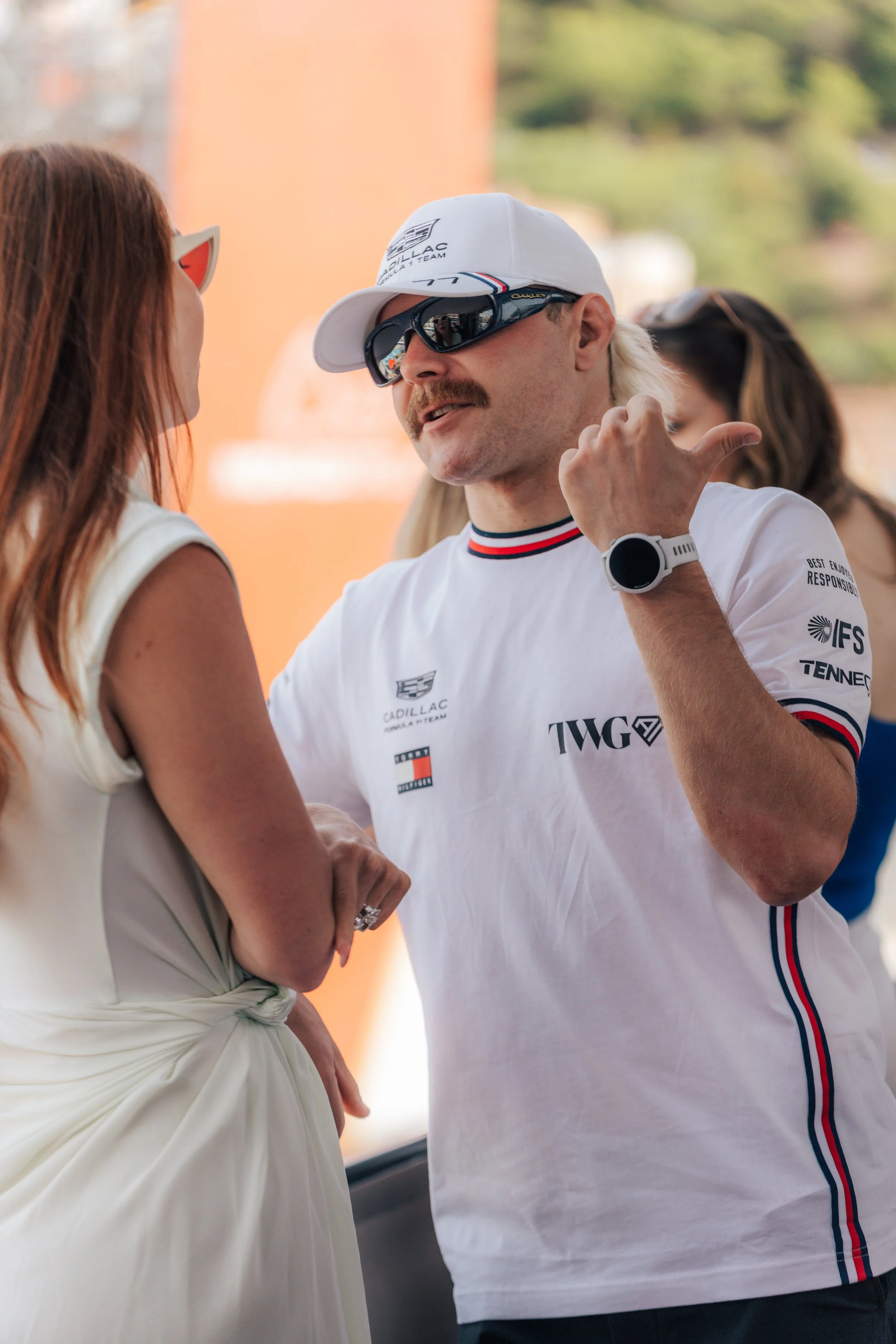 A man with a white cap, sunglasses, and a white racing shirt talking to a woman with long brown hair and sunglasses, outdoors with a blurred background.