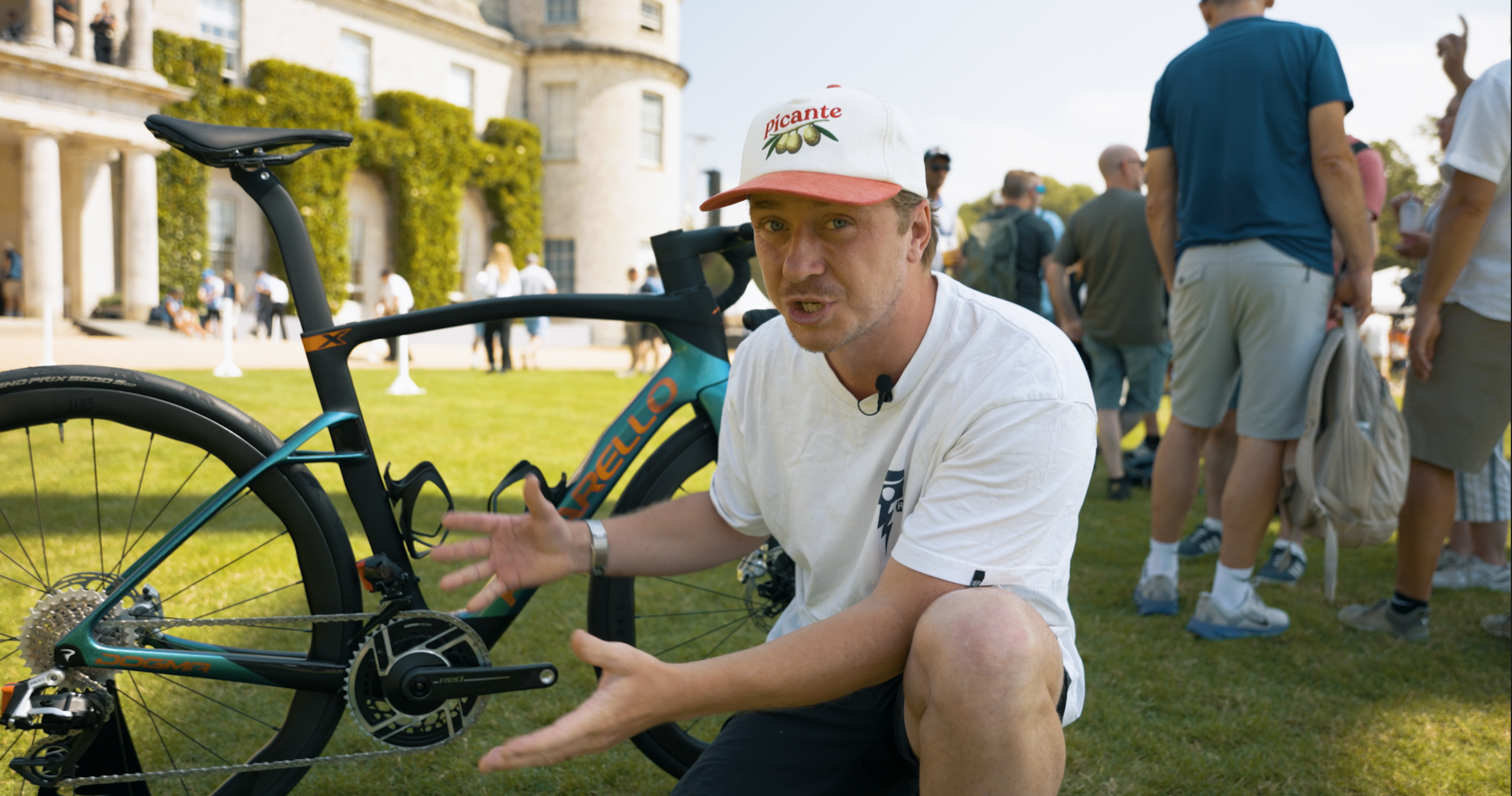 A man kneeling on the grass, pointing to a teal and black Cannondale bike, with a historic castle or mansion in the background. Several people are gathered around, some standing and talking, in a sunny outdoor setting.