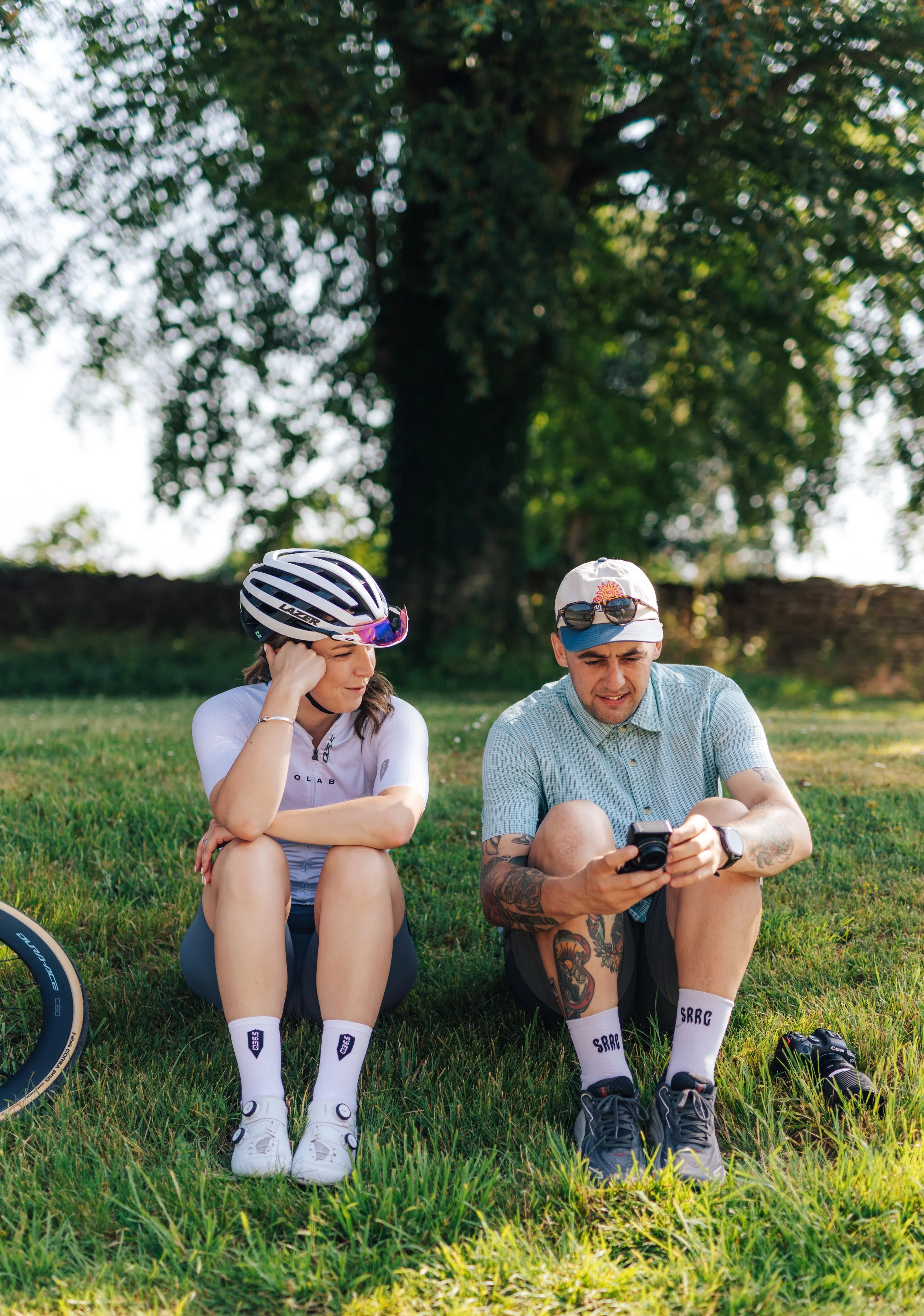 A woman and a man sitting on grass in a park, looking at a camera and a phone, with trees in the background.