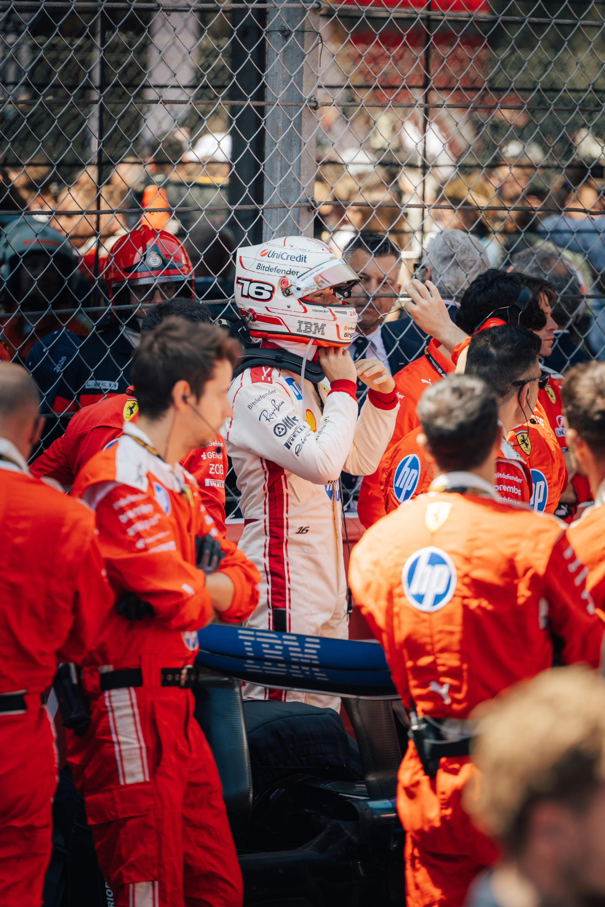 A group of Formula 1 teams and drivers at a racetrack, with some members adjusting gear and helmets, behind a safety barrier.