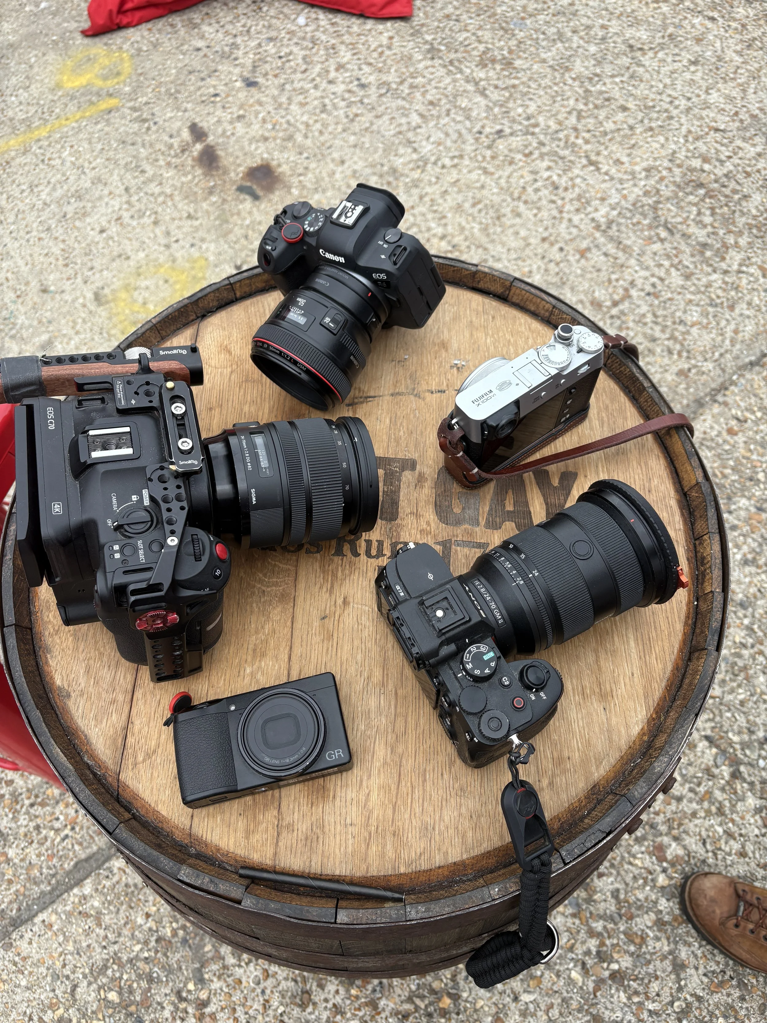 Several cameras and camera accessories, including a Canon EOS camera with a lens, a smaller Sony camera, and a FujiFilm camera, are arranged on a round wooden table outdoors.