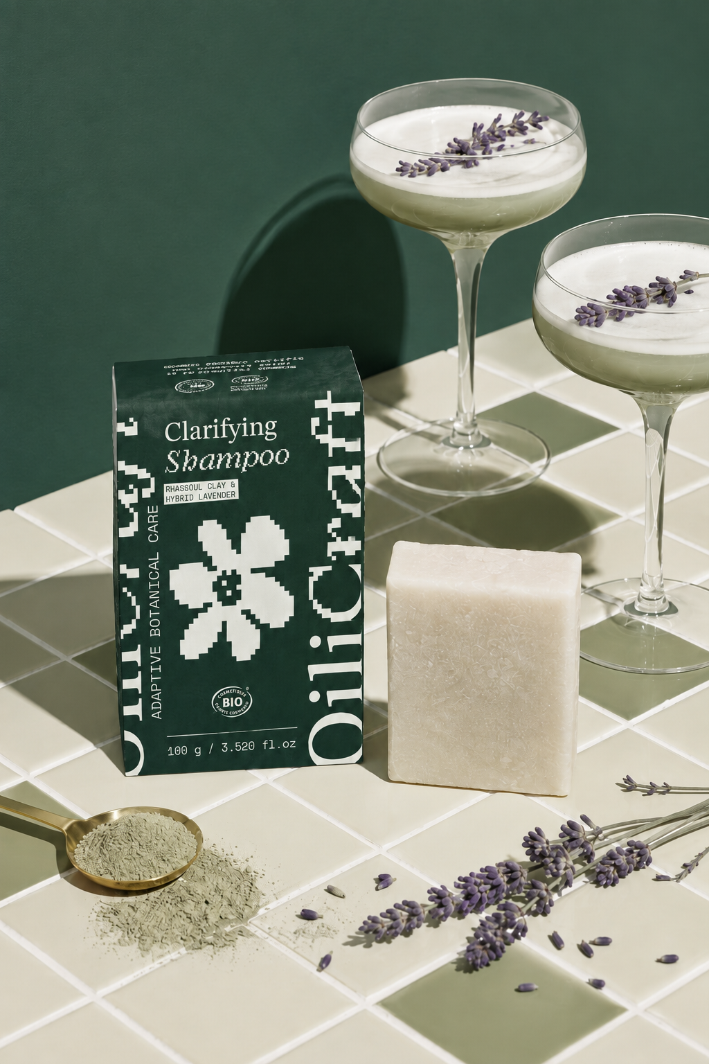 Deux coupes à cocktail avec lavande en décoration, un sachet de shampoing clarifiant à base de lavande, un savon en bloc, une cuillère avec de la poudre de lavande, et des brins de lavande sur une surface en carreaux blancs.