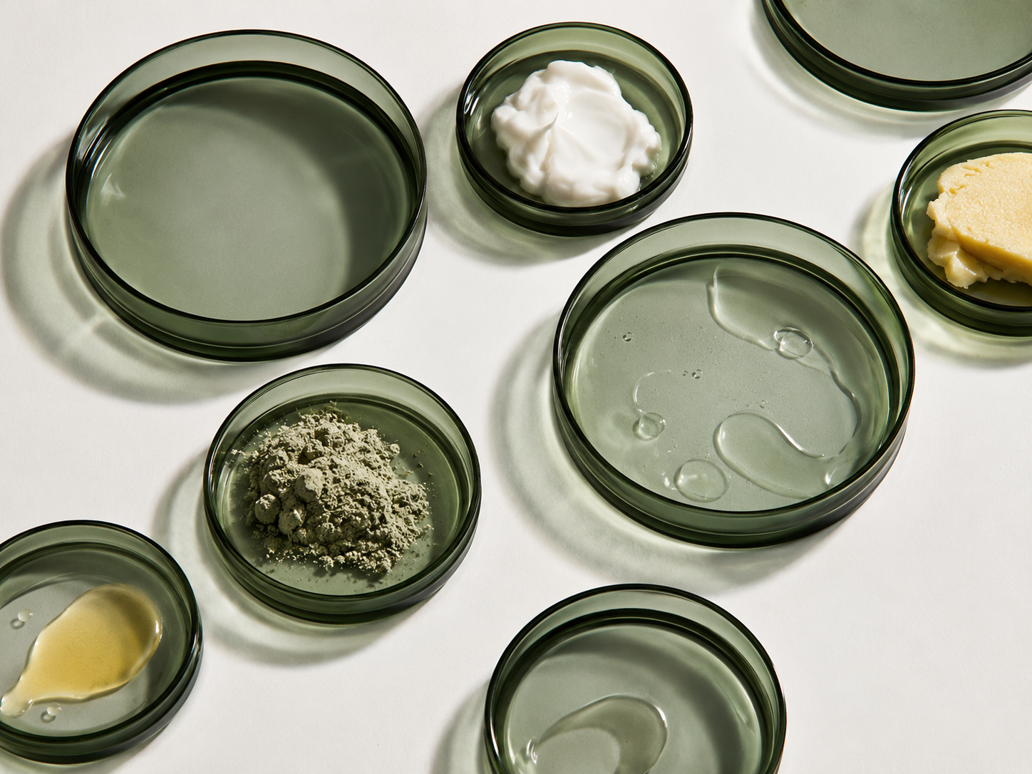 Différents contenants en verre avec diverses substances, y compris une crème blanche, de la pâte, de la poudre, et des liquides, sur une surface blanche.