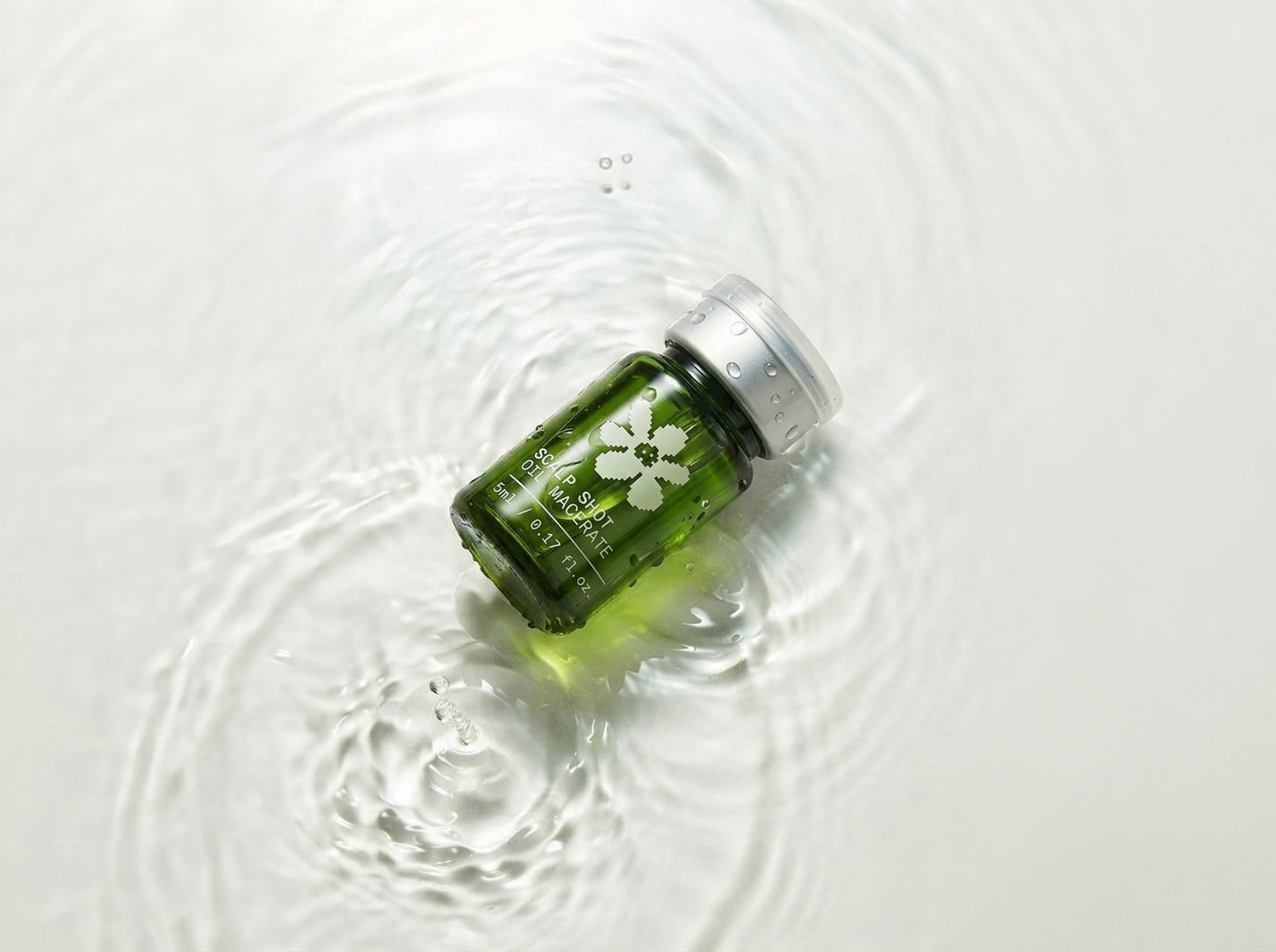 Une petite bouteille en verre de couleur verte flottant dans l'eau, portant une étiquette indiquant 'Scalp Shot Macérate' et une fleur blanche stylisée, avec des gouttes d'eau sur le goulot et une surface d'eau calme.