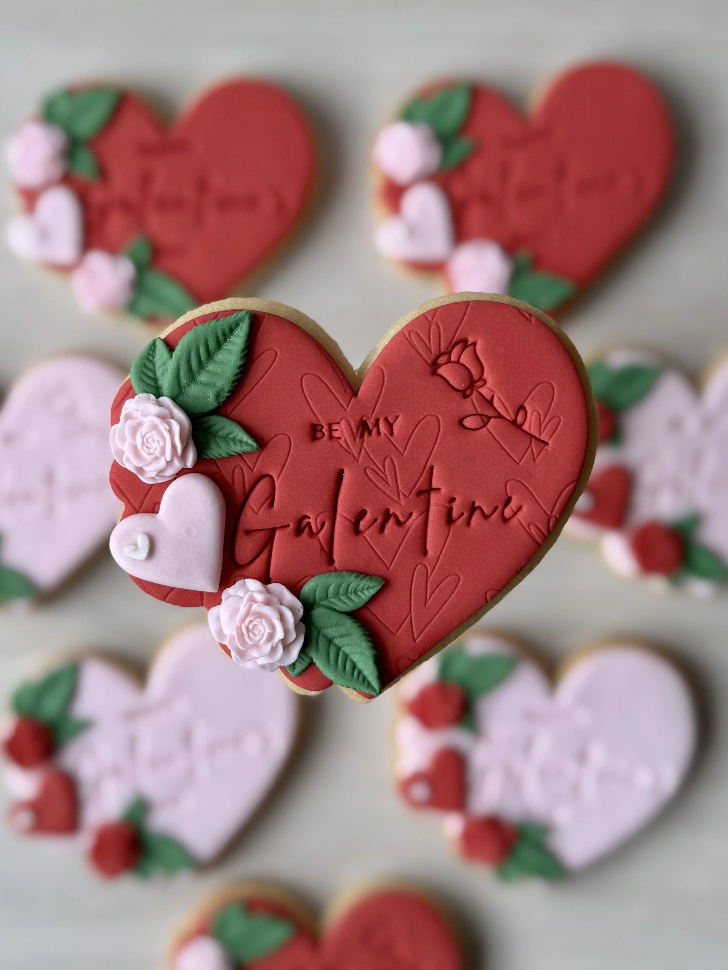 Decorated heart-shaped cookies with pink and red fondant icing, green leaves, white and pink roses, and the words 'BE MY Galentine' inscribed