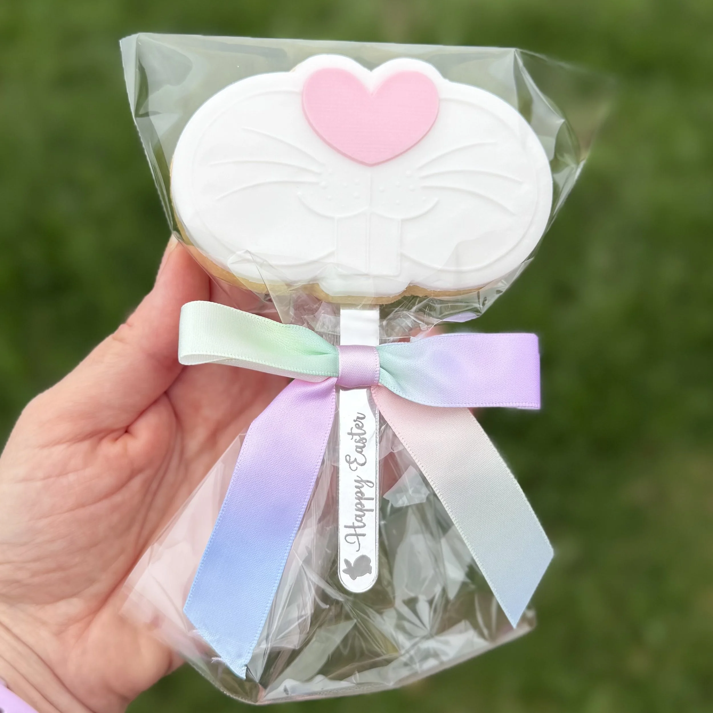 A person holding a decorated cookie with an Easter bunny nose, with a pink heart and tied with a rainbow ribbon