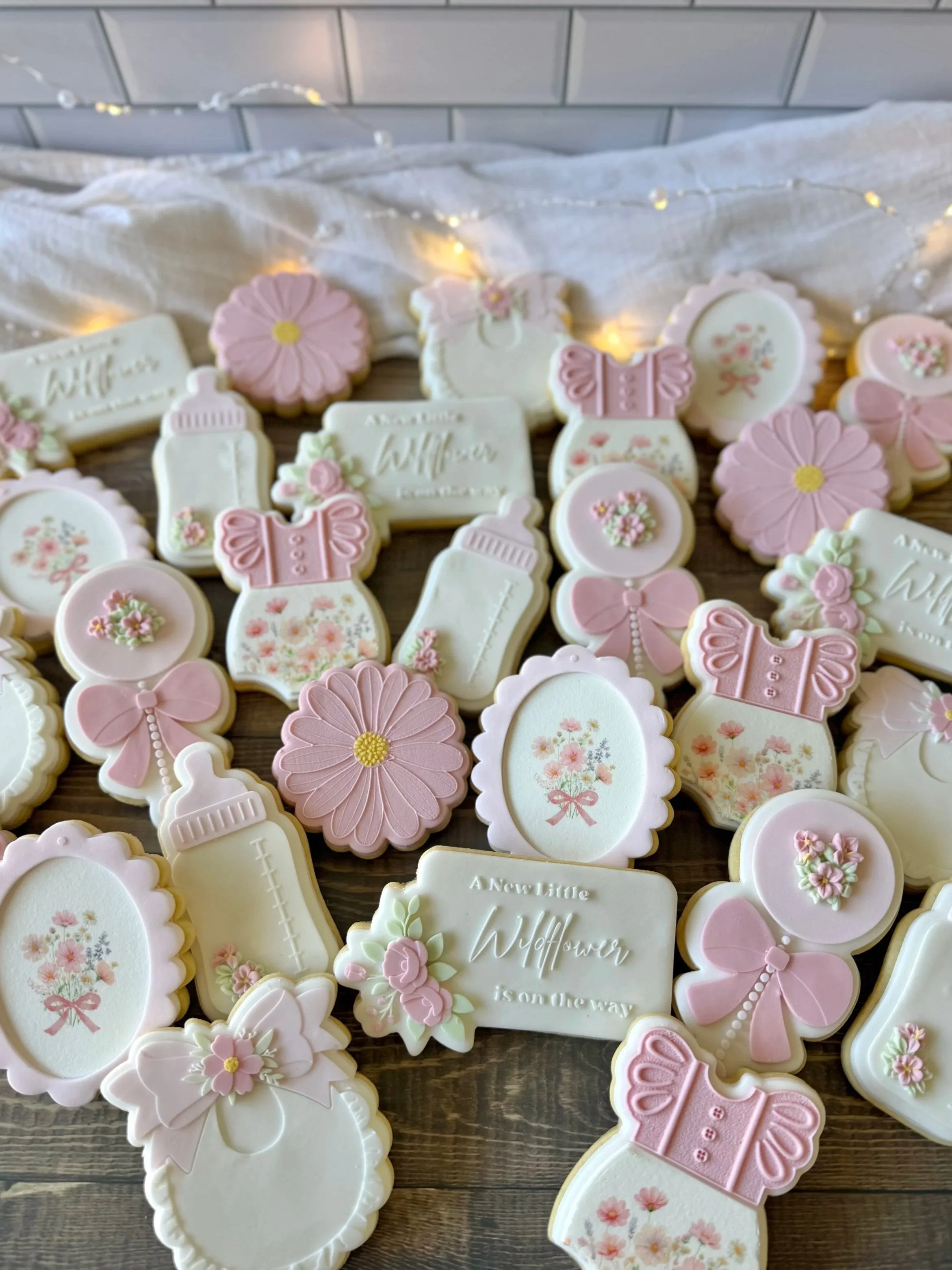 Assorted pastel-colored cookies with floral, bow, and baby bottle designs on a wooden surface, with a white draped fabric background and string lights