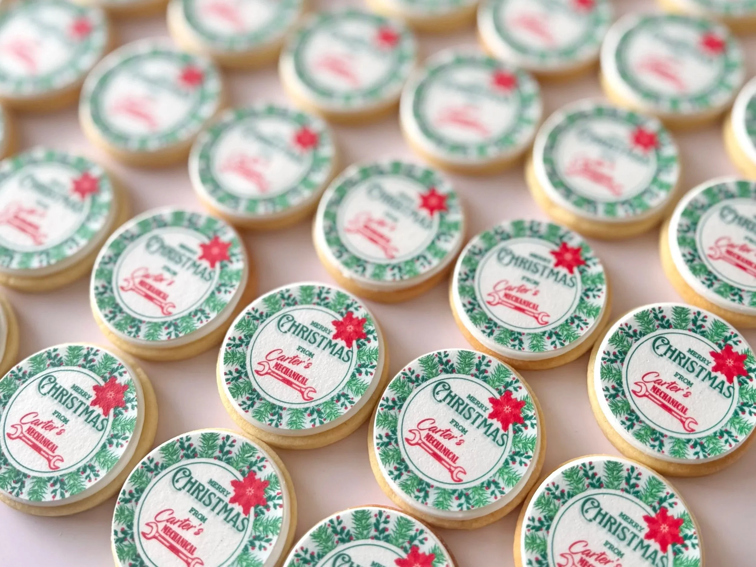 Close-up of multiple round cookies with Christmas-themed edible printed decorations, featuring pine branches and a red poinsettia flower, with the text "Merry Christmas from Carter's Mechanical".