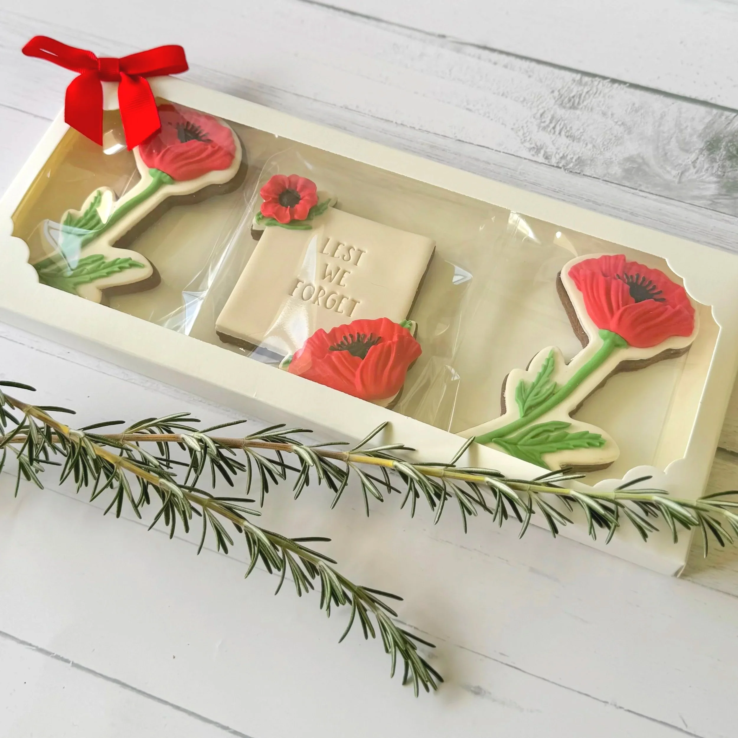 Close-up photo of decorated ANZAC cookies with floral designs, a gift box, and sprigs of rosemary on a white wooden surface