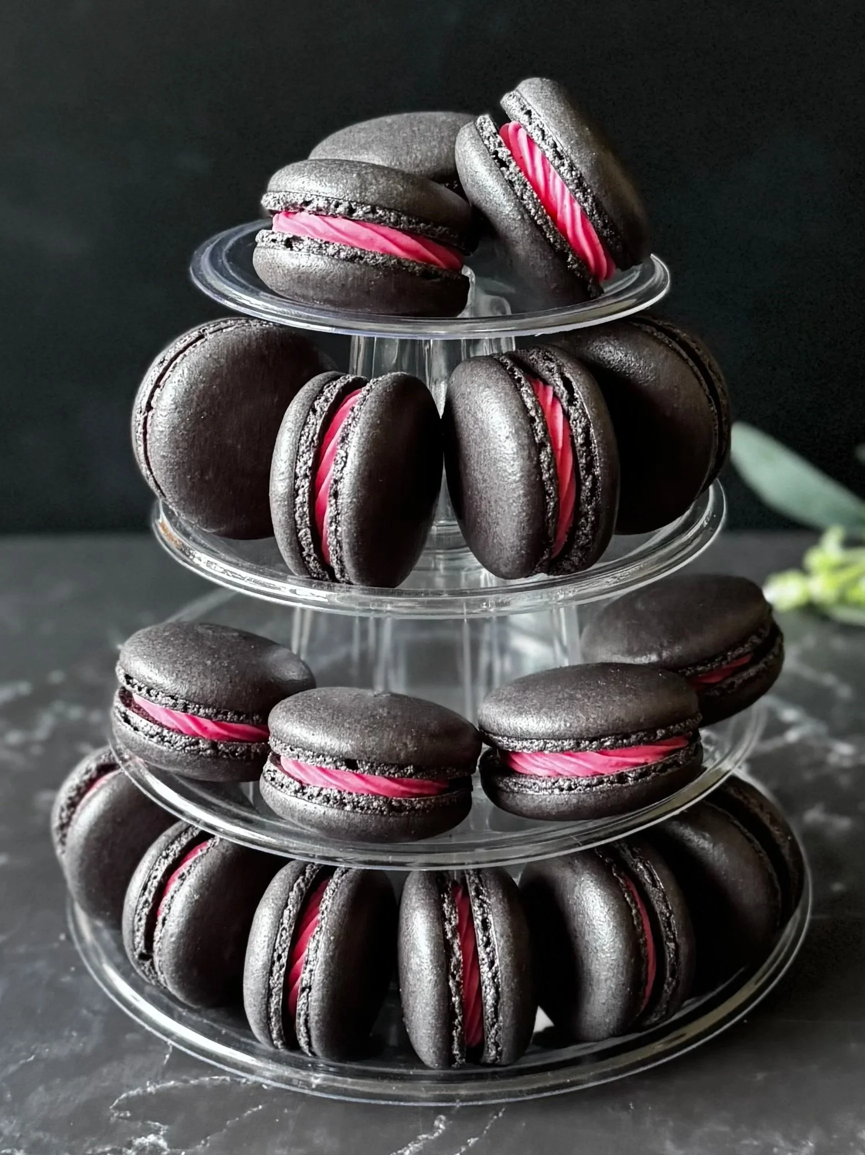 Stacked black macarons with pink filling on a clear multi-tiered serving tray against a dark background
