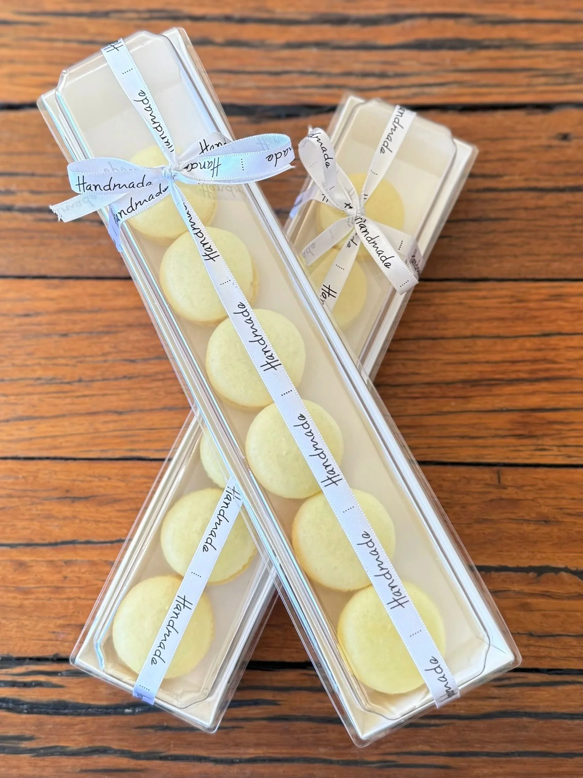 Two clear plastic boxes of yellow macarons tied with white ribbons that say 'Handmade'