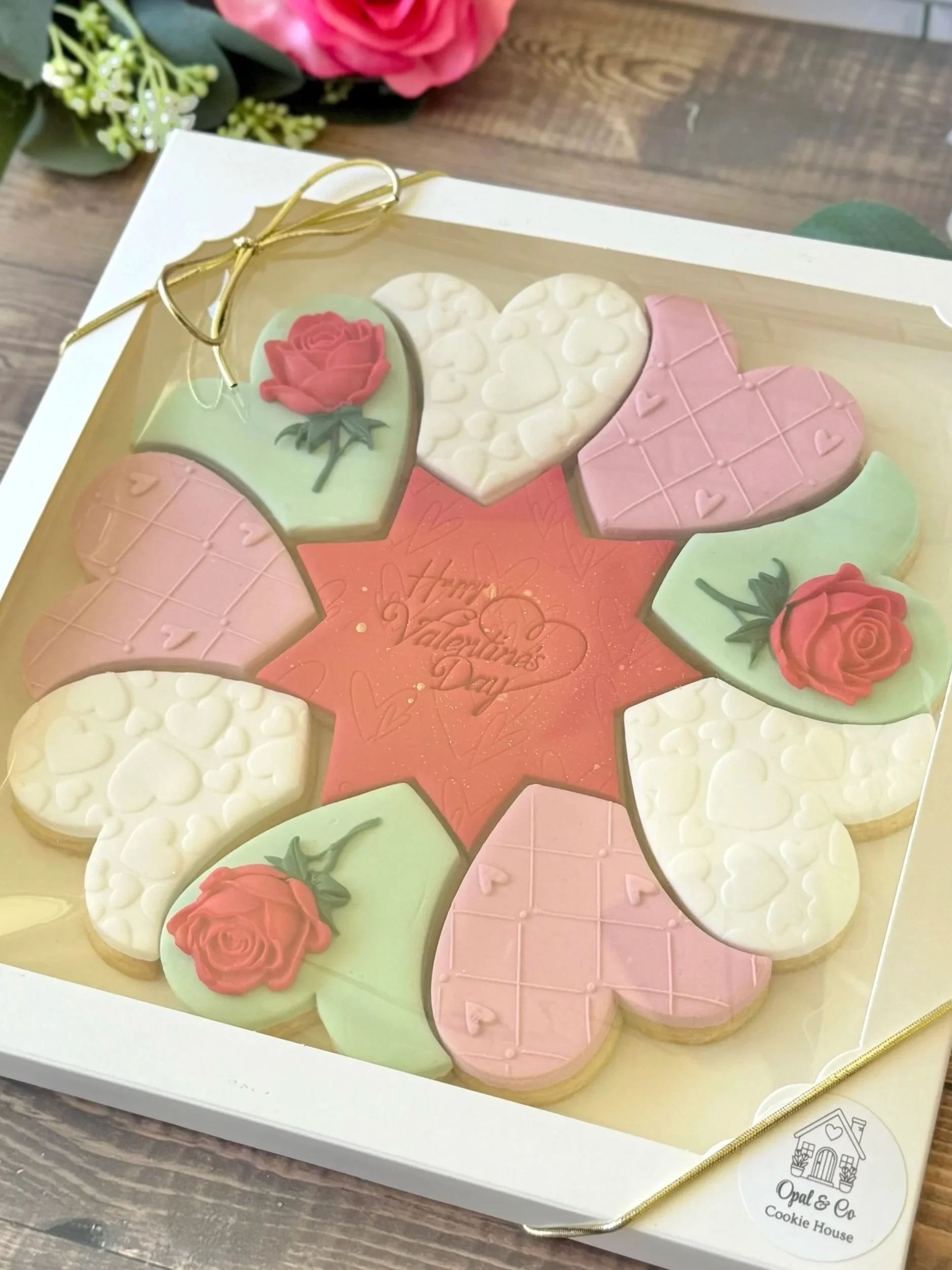 A box of decorated cookies with floral and love-themed designs, some cookies have pink and green icing with roses, and others are white with embossed hearts, all arranged in a star shape for Valentine's Day