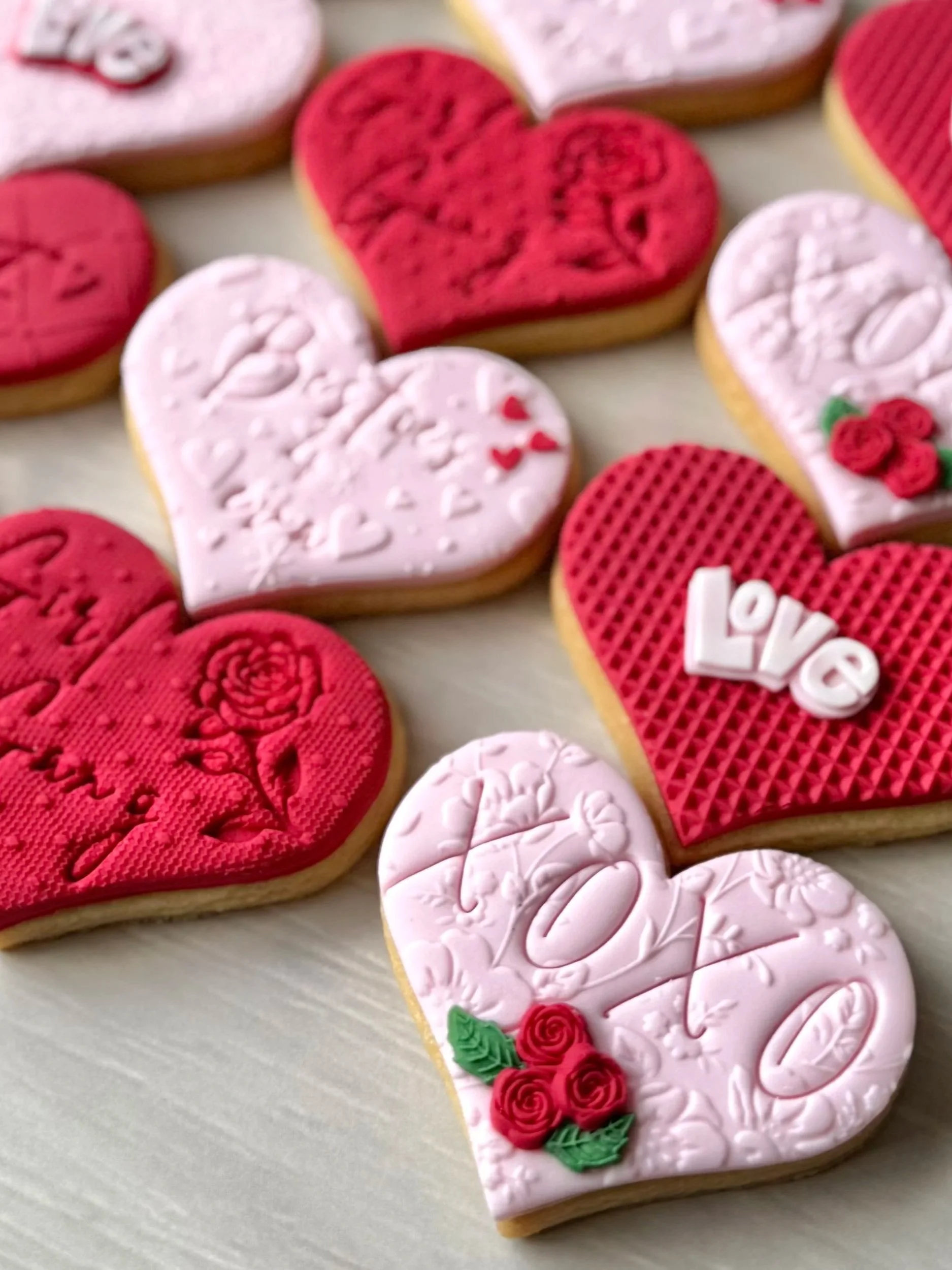 Decorative Valentine's Day cookies in heart shapes with pink and red icing, some with roses, the word 'Love', and embossed patterns.