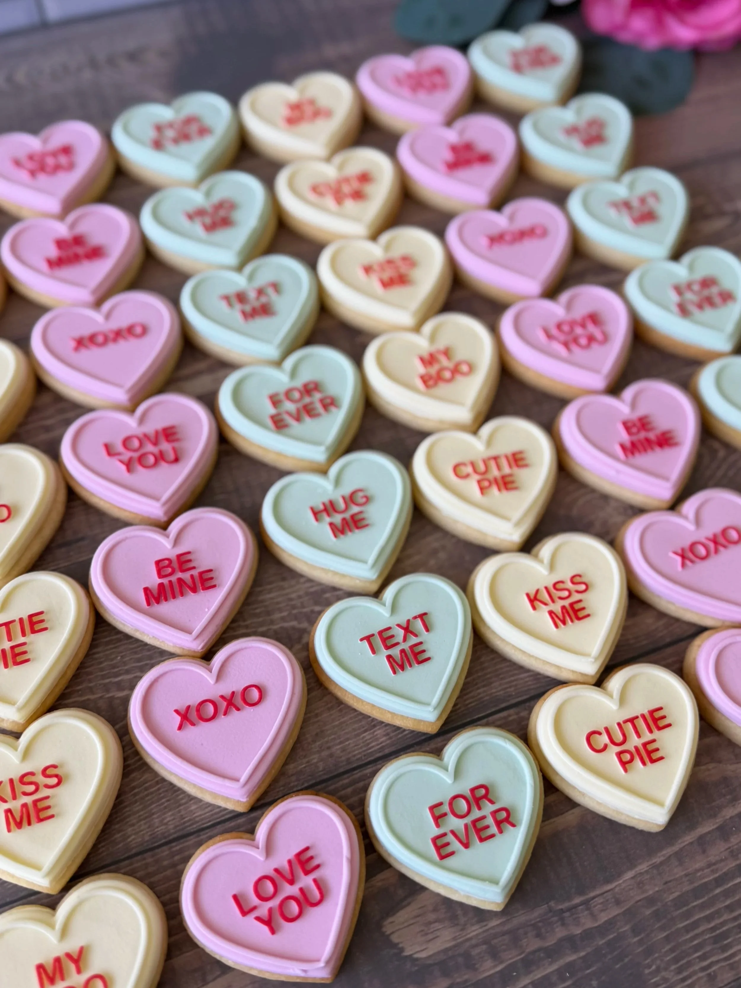 Colourful heart-shaped sugar cookies with romantic messages like "XOXO," "LOVE YOU," "BE MINE," "KISS ME," "TEXT ME," "HUG ME," and "FOR EVER"