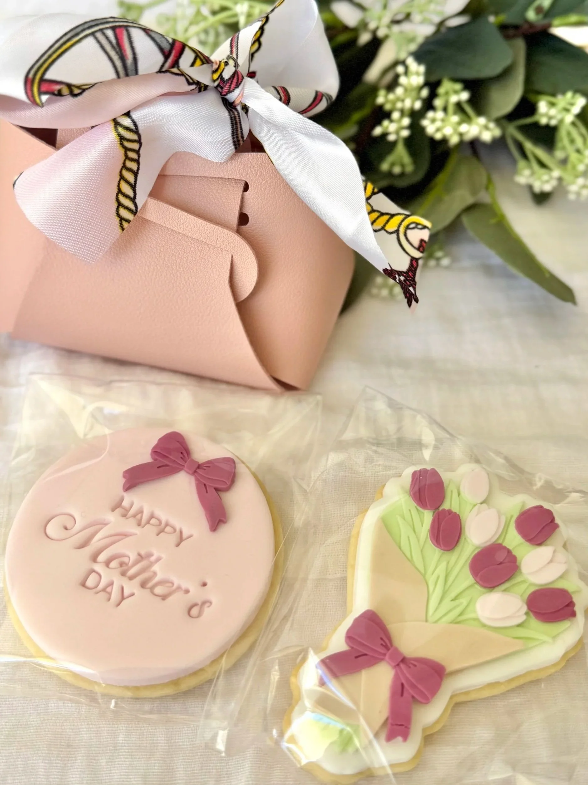 Decorative cookies with floral designs, a pink cookie with a pink bow and the words 'Happy Mother’s Day,' and a pink mini handbag with a floral wreath in the background