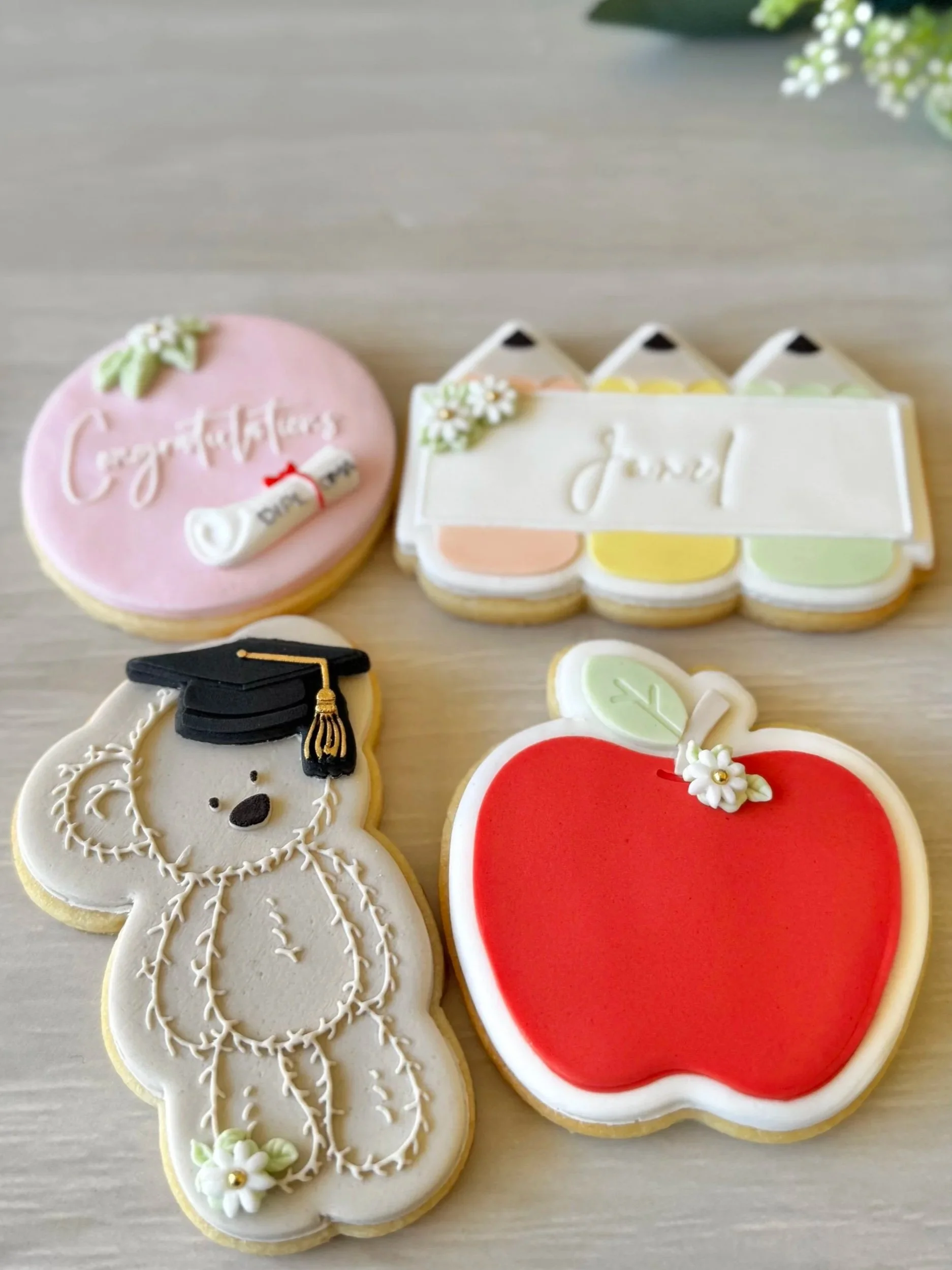 Decorated fondant sugar cookies for a graduation celebration, including a bear with a graduation cap, a diploma scroll, a 'congratulations' cookie, a red apple with a leaf and flower, and colourful pencils personalised with the graduate's name