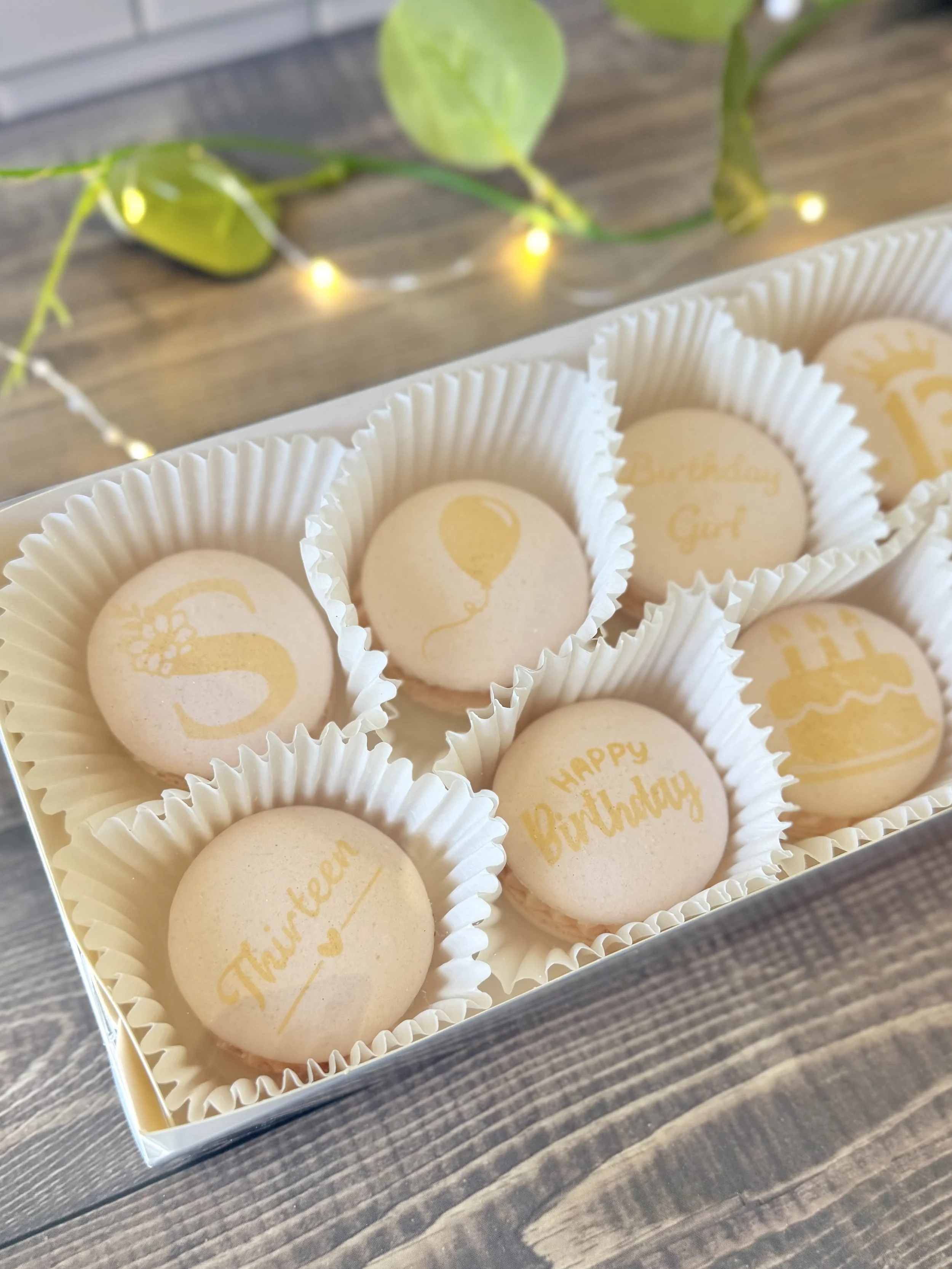 A box of beige birthday macarons decorated with gold writing and various images created by airbrushing custom stencils. The macarons are in white paper cups and are arranged neatly inside the box