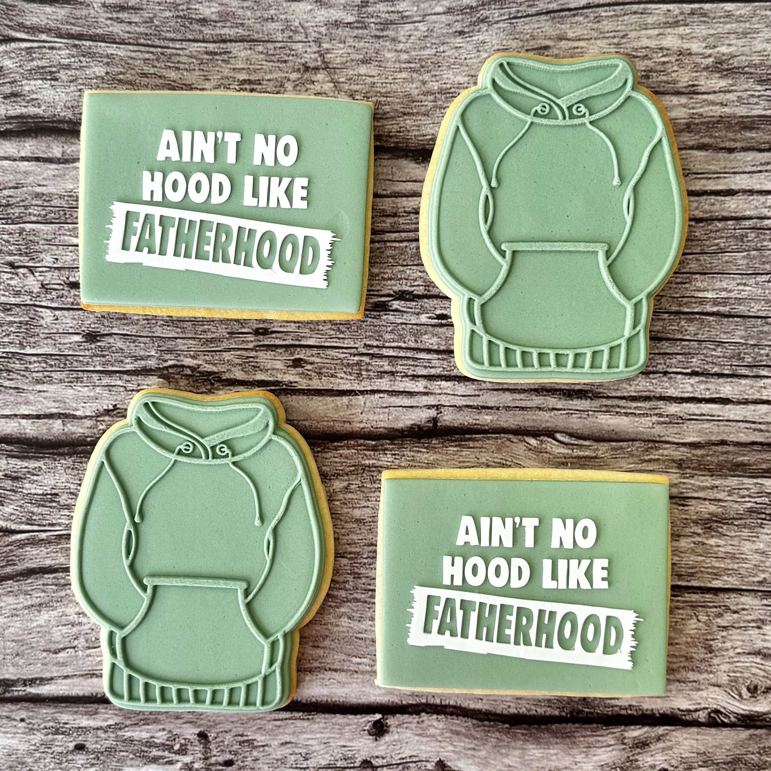 Decorated cookies on a wooden surface; one cookie has the phrase "Ain't no hood like Fatherhood" written on it, and the other is shaped like a hoodie with green fondant