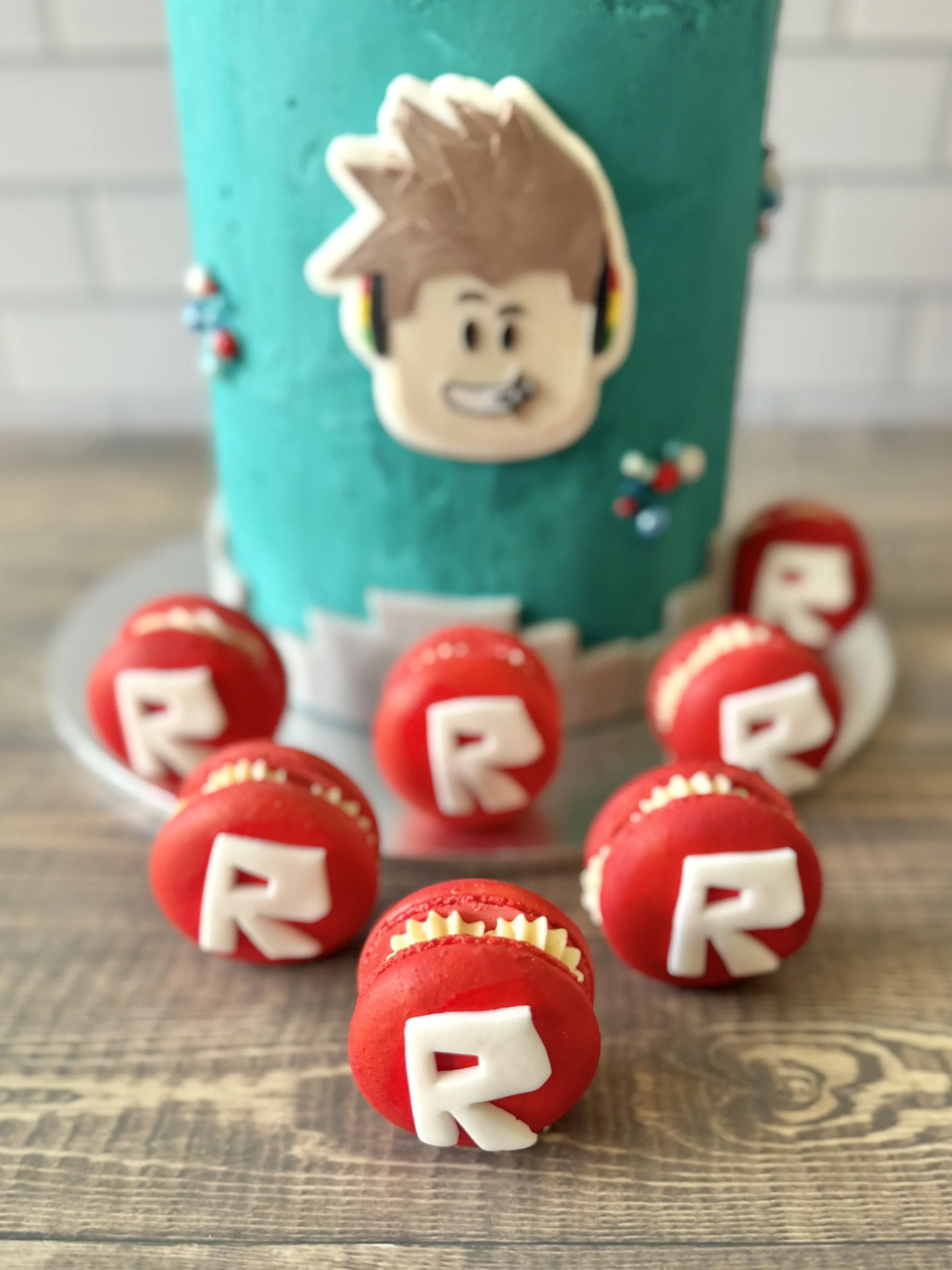 Red macarons with white letter 'R' on top, arranged in front of a teal-covered cake with a cartoon gaming face and headphones