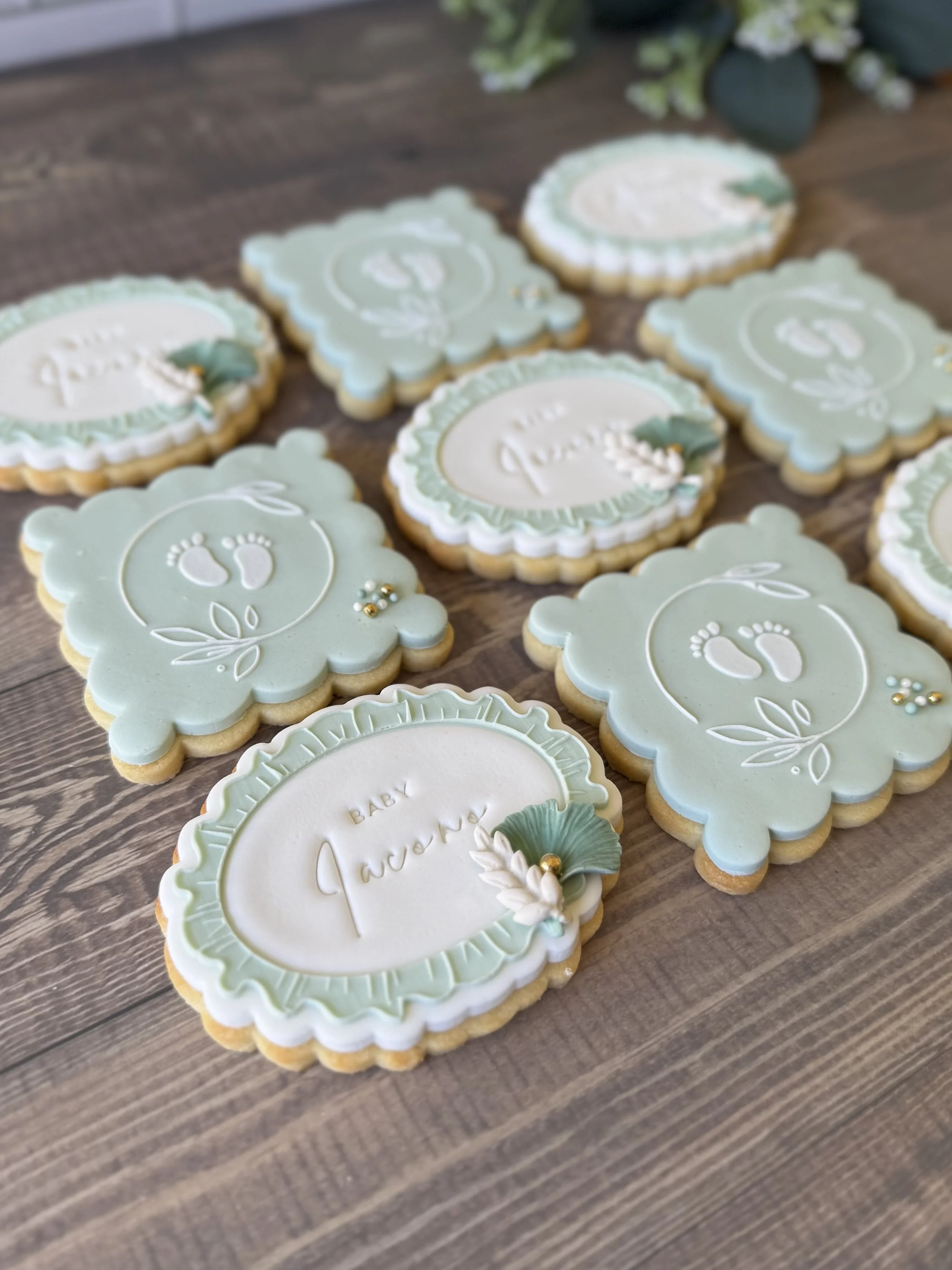 Cookies with pale green and white fondant, decorated with baby footprints and floral designs, arranged on a wooden surface