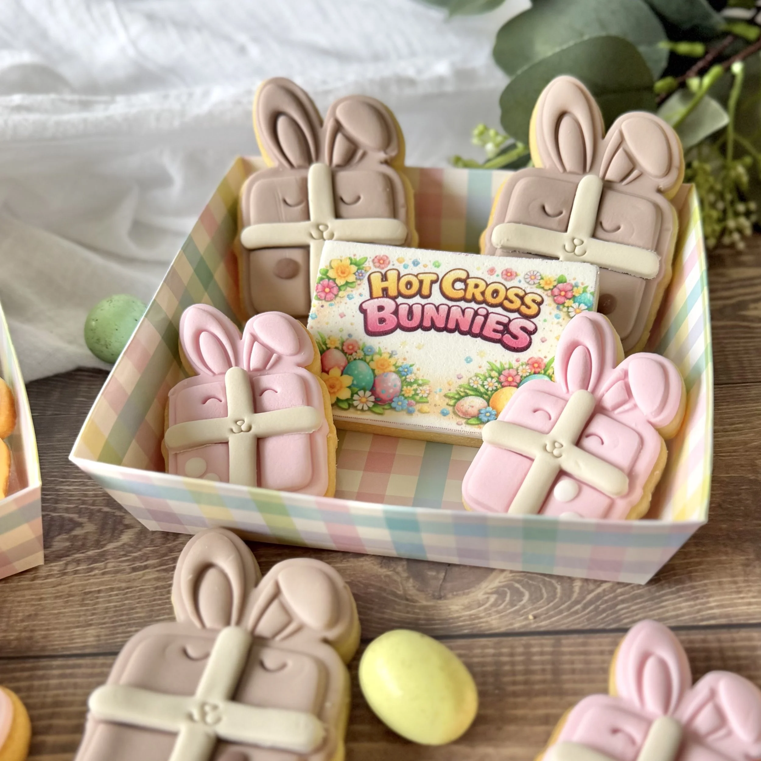 A collection of Easter-themed cookies including bunny-shaped cookies with pink and white fondant, and a sign that says "Hot Cross Bunnies," inside a pastel-coloured checkered box