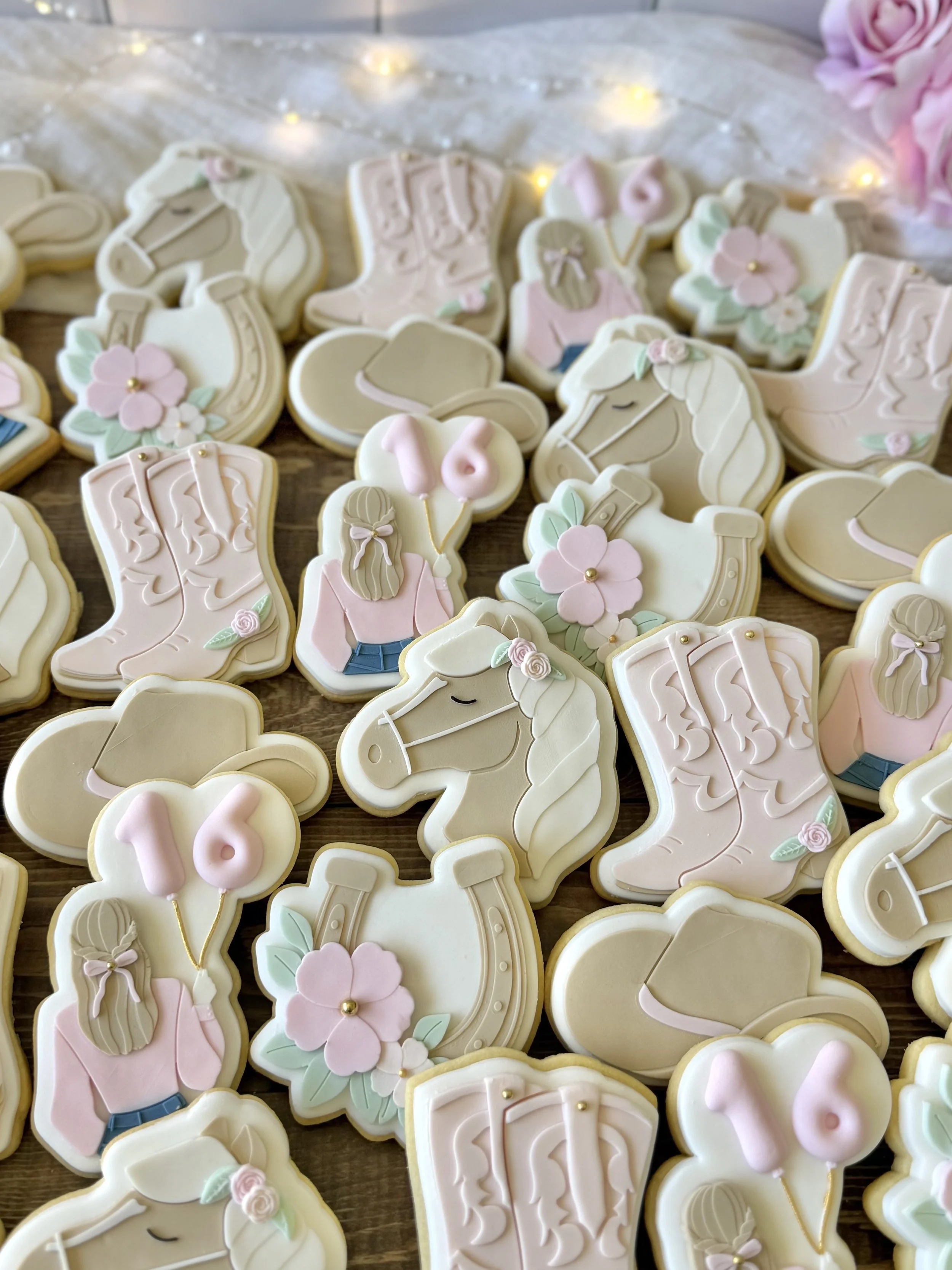 Western-themed 16th birthday cookies featuring horses, cowgirl boots, cowboy hats, floral horseshoes and personalised number 16 cookies in soft pink, cream and brown