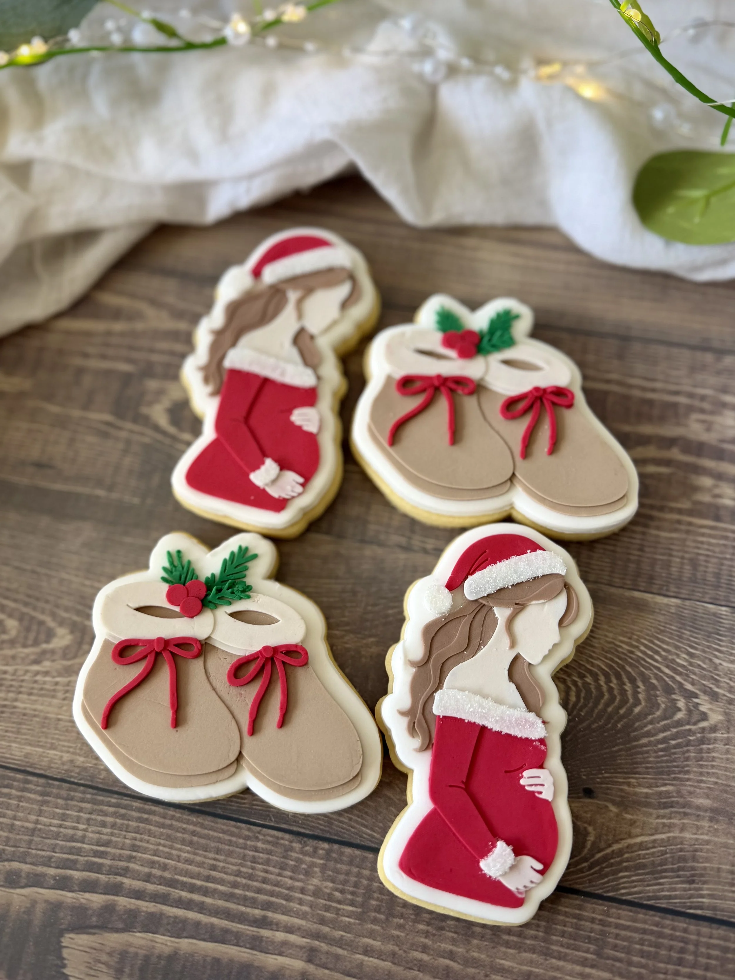 Handcrafted Christmas-themed sugar cookies featuring a beautiful mum-to-be dressed in red with a Santa hat, paired with adorable baby booties decorated with festive bows, holly and layered fondant details. Perfect for Christmas pregnancy announcement
