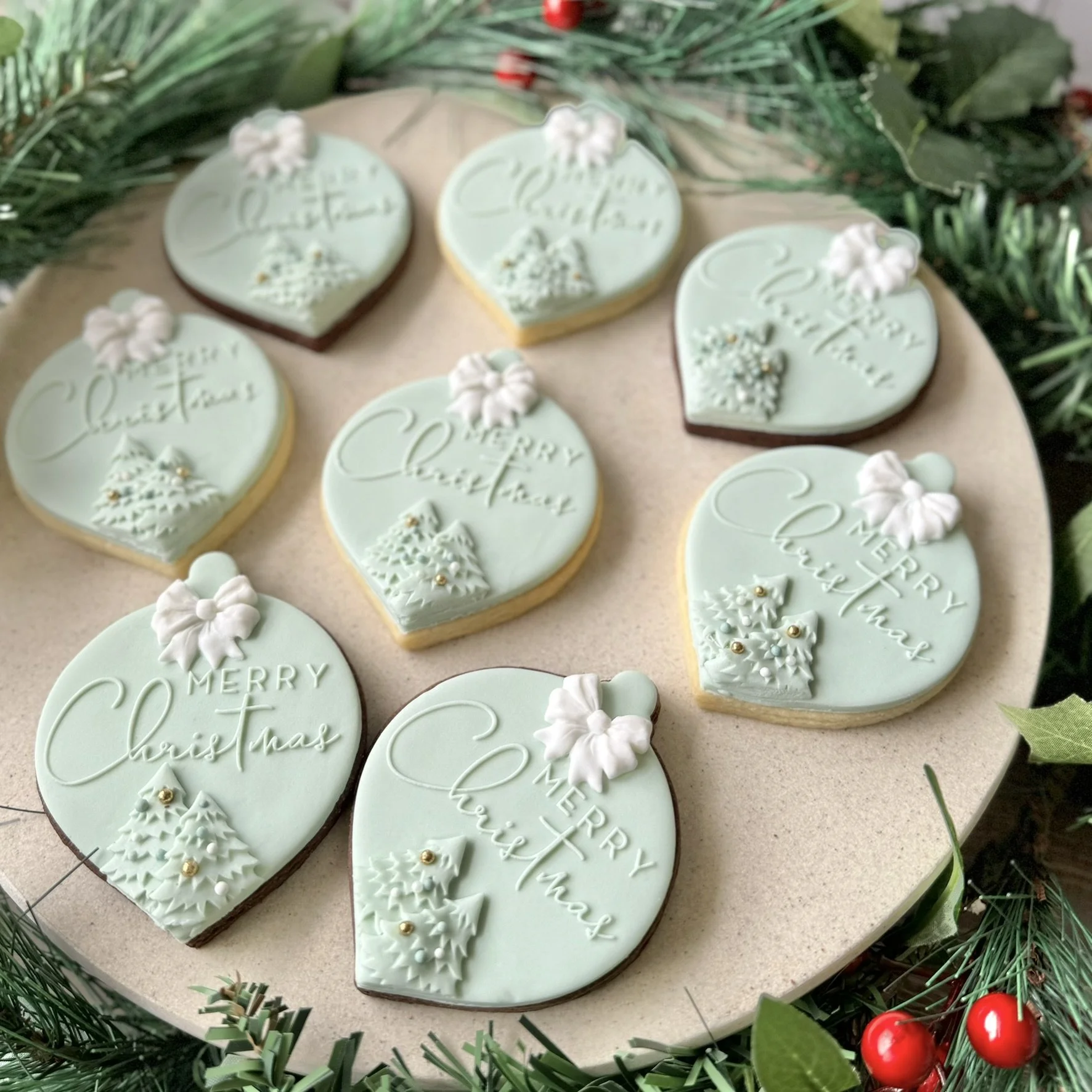 Decorative Christmas cookies with pine tree designs, and holiday messages, arranged on a round tray surrounded by festive greenery and red berries