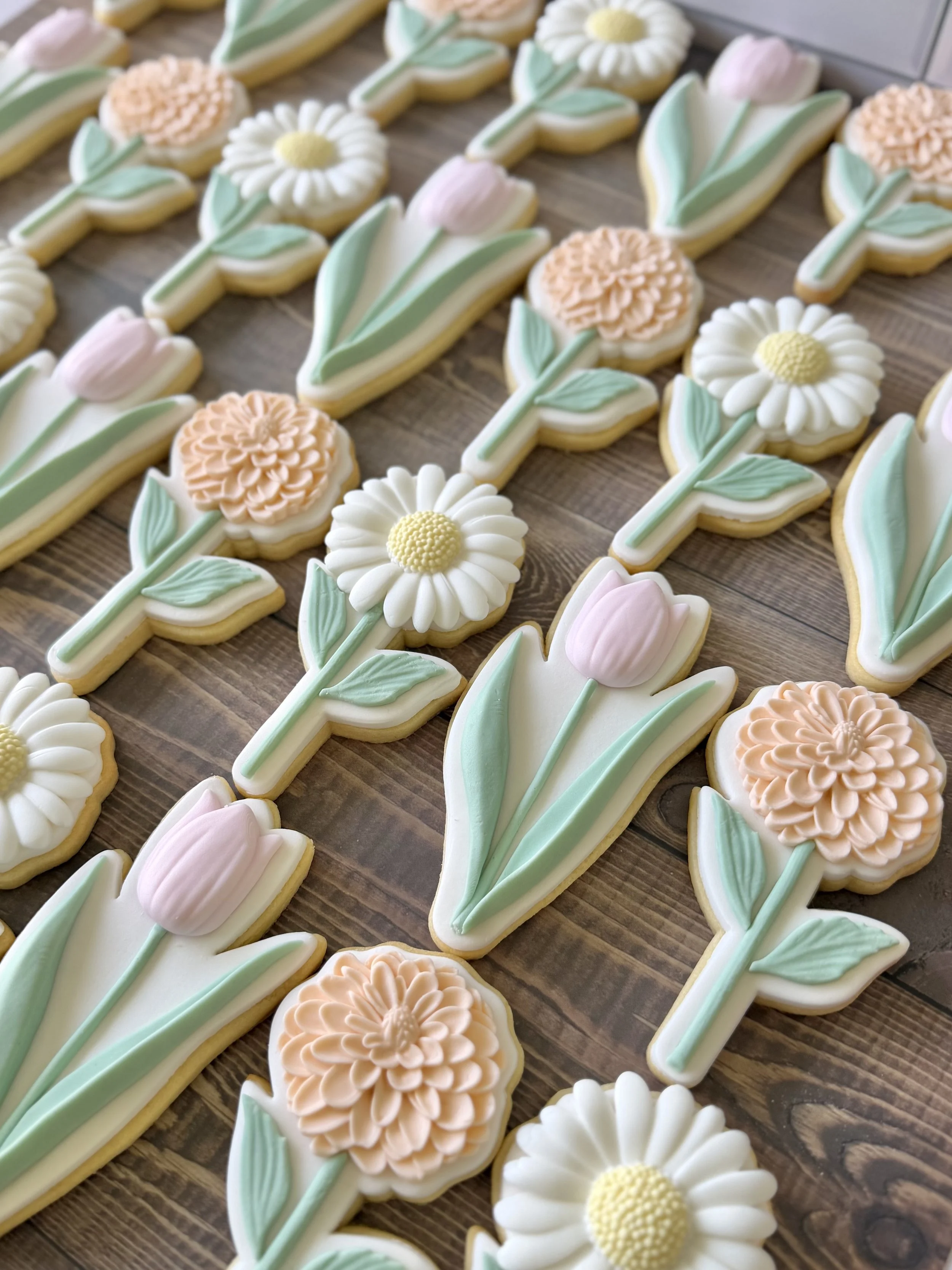 Assortment of flower-shaped cookies decorated with pastel-colored icing, including daisies, tulips, and dahlia designs, arranged on a wooden surface.