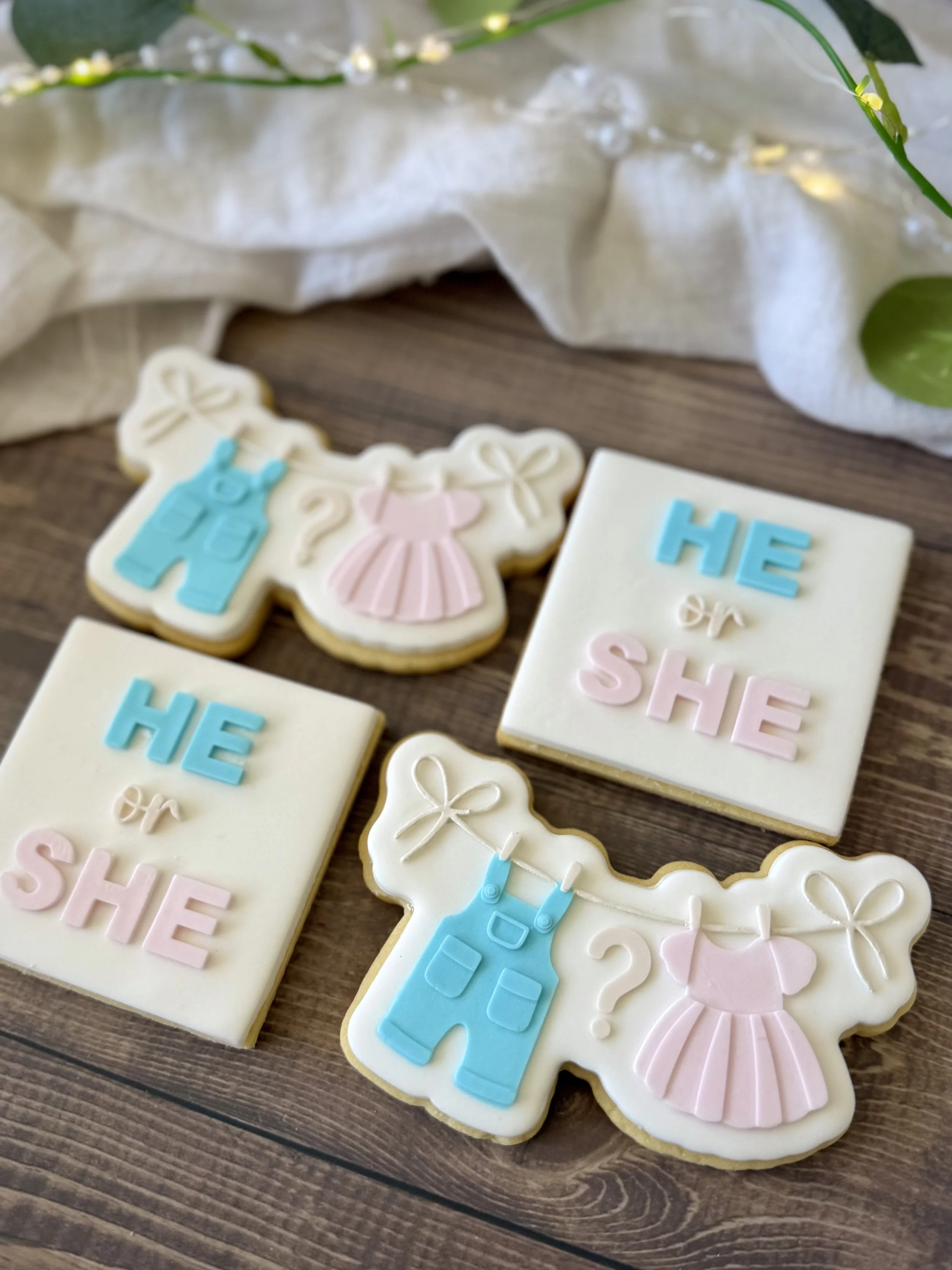Handmade gender reveal sugar cookies featuring He or She lettering and adorable baby clothes hanging on a line. Decorated with layered fondant in pastel pink, blue and white.