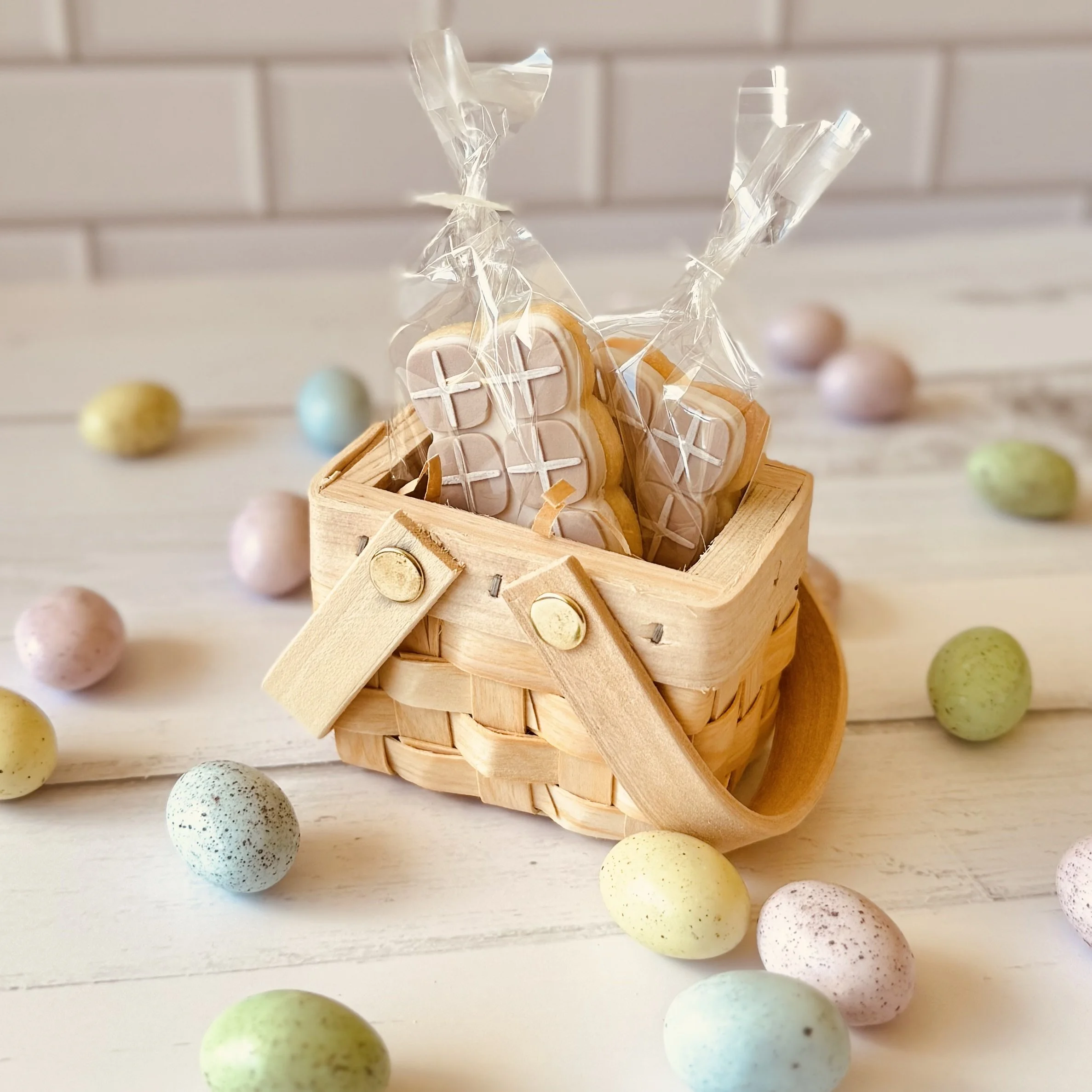 Colourful speckled candy eggs arranged around a small wooden basket with clear plastic wrapping, set on a white surface with a white tiled background, featuring mini hot cross buns